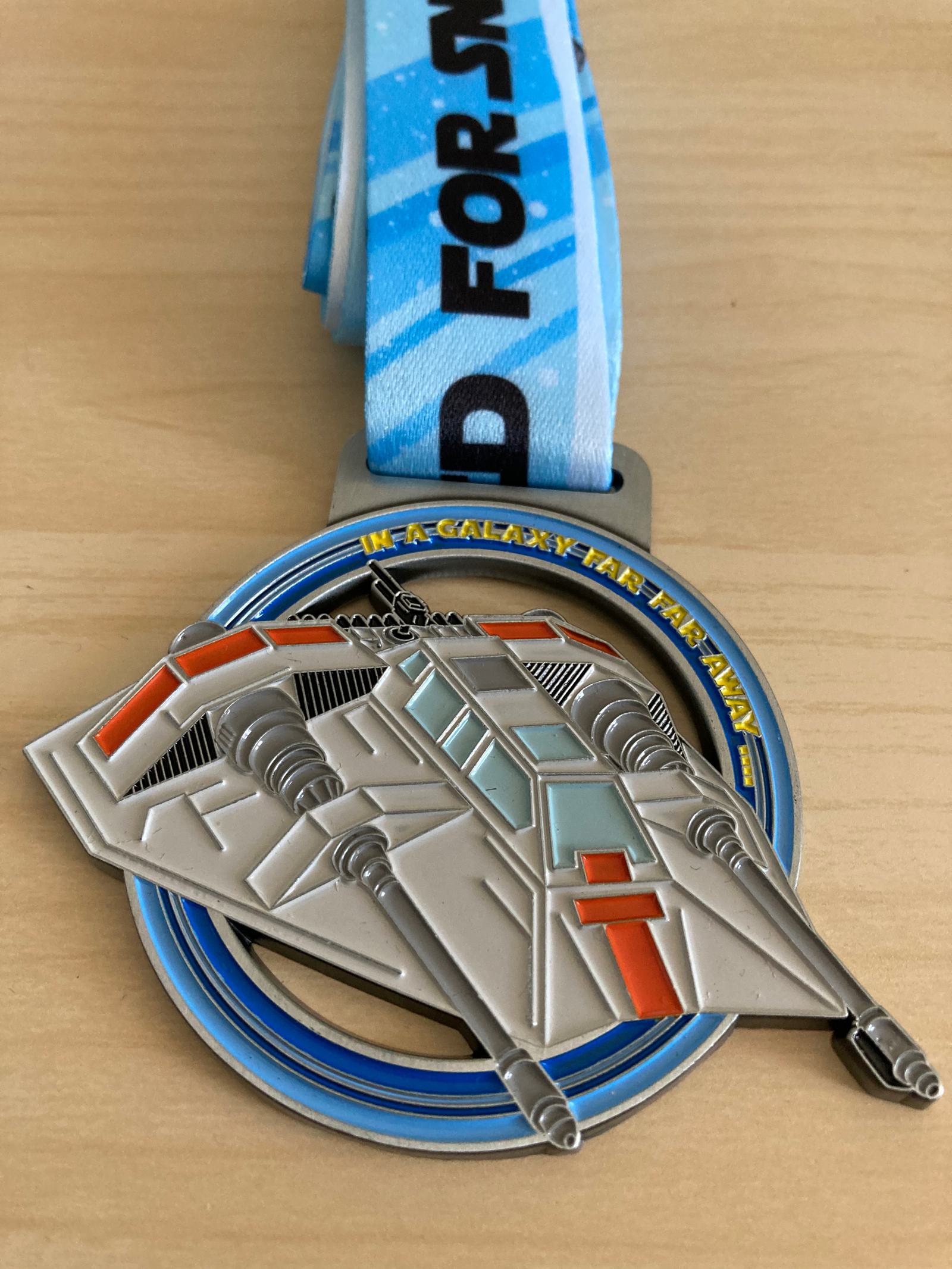 A medal with a blue ribbon featuring a spaceship design. The medal depicts a detailed spaceship with orange and gray accents. The ribbon has the text "I RAN A GALAXY FAR FAR, AWAY" printed on it. The background is a wooden surface.