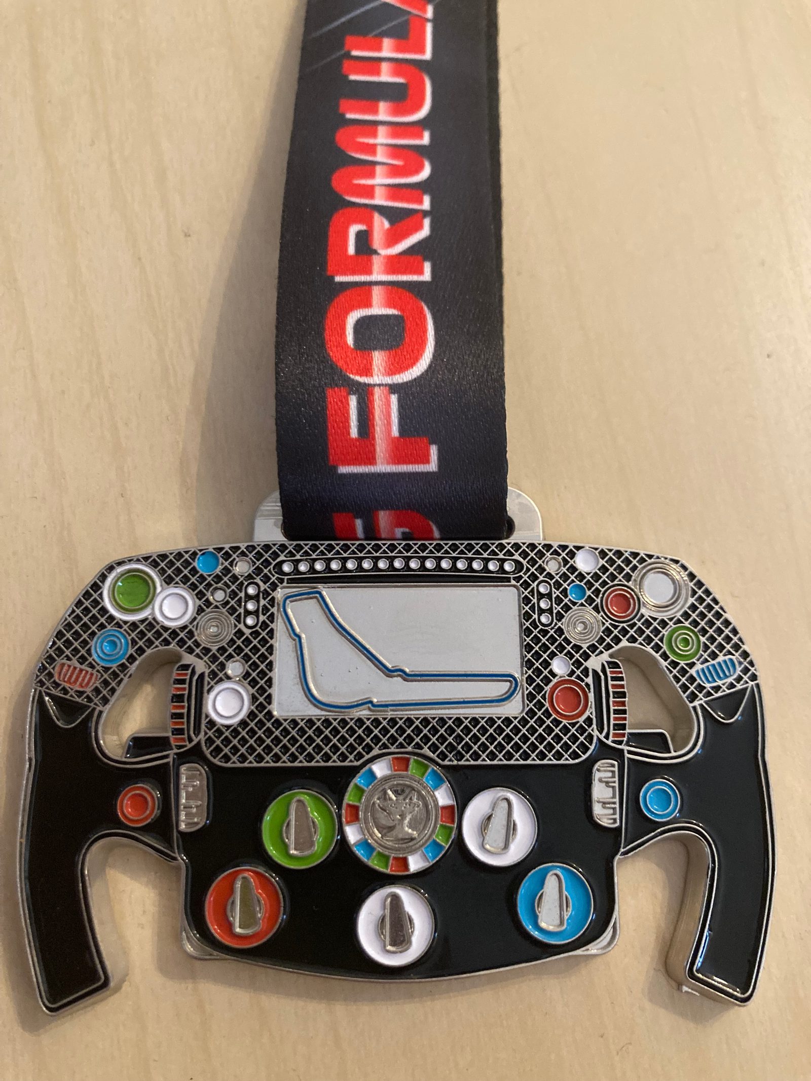 A medal shaped like a Formula 1 steering wheel, with simulated control buttons and knobs in various colors. The ribbon attached to the medal is black with the word "FORMULA" in red font. The medal's center features a race track outline in blue.