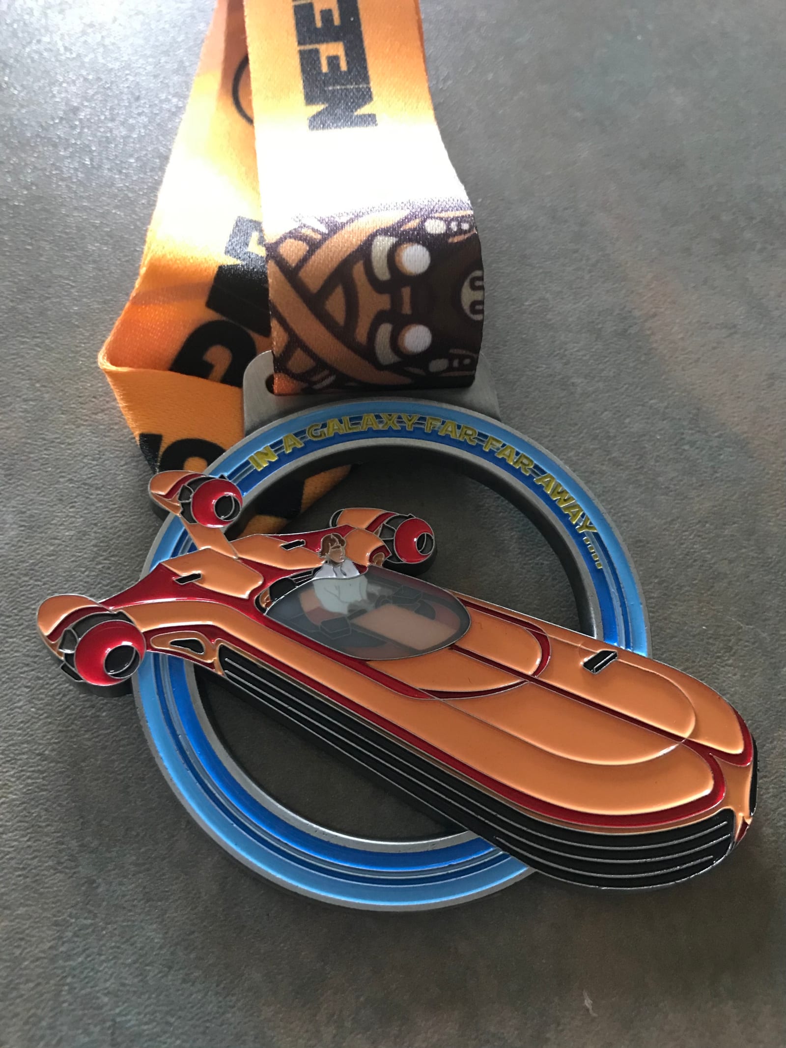 A colorful race medal features an orange futuristic hovercraft with a pilot, attached to an orange and blue ribbon. The text on the blue ring of the medal reads, "In a galaxy far far away.