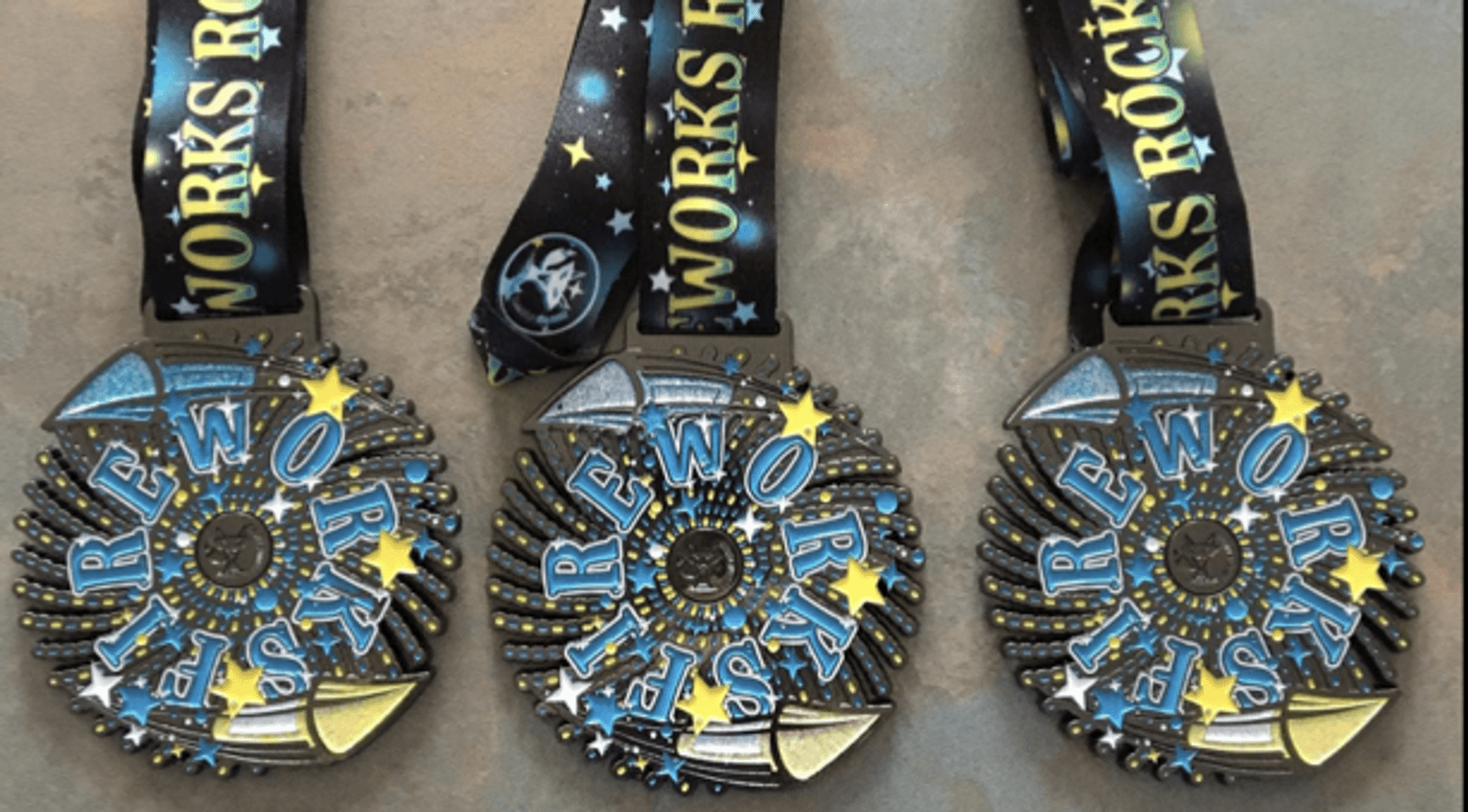 Three "Fireworks Rock!" medals with black ribbons adorned with stars and lettering that says "Fireworks Rock!" The medals are colorful, featuring blue, yellow, and white designs with star accents arranged in a circular pattern.
