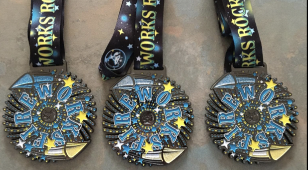 Three "Fireworks Rock!" medals with black ribbons adorned with stars and lettering that says "Fireworks Rock!" The medals are colorful, featuring blue, yellow, and white designs with star accents arranged in a circular pattern.