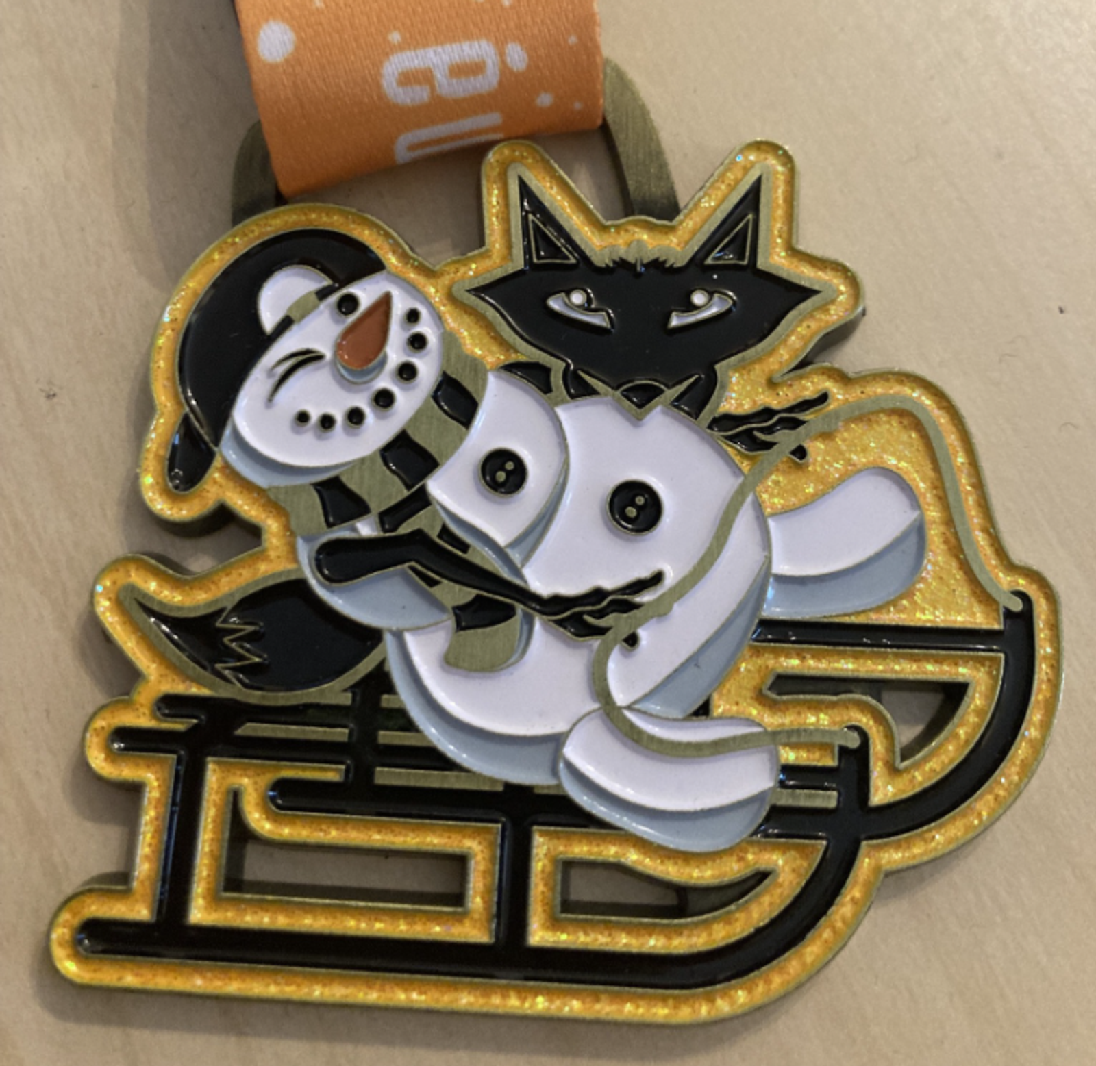 A festive pin featuring a joyful snowman and a black cat on a golden sled. The snowman, wearing a hat and scarf, smiles with outstretched arms. The cat, with pointed ears and a content expression, sits behind the snowman. The background is a light surface.