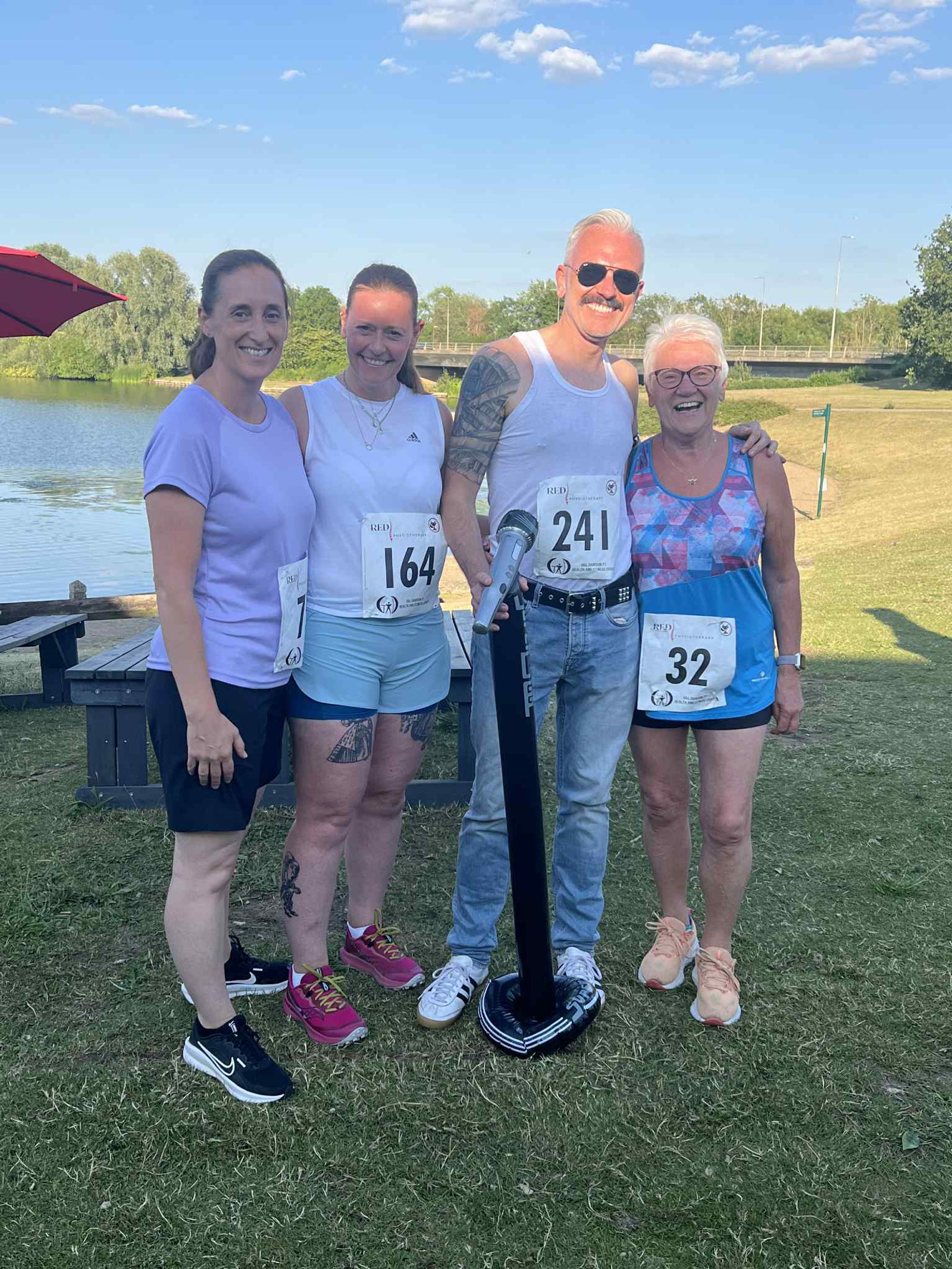 Four smiling people with race bibs stand together on grass by a lake, under a sunny blue sky. One man holds a large microphone. The group appears happy and relaxed after an outdoor event.