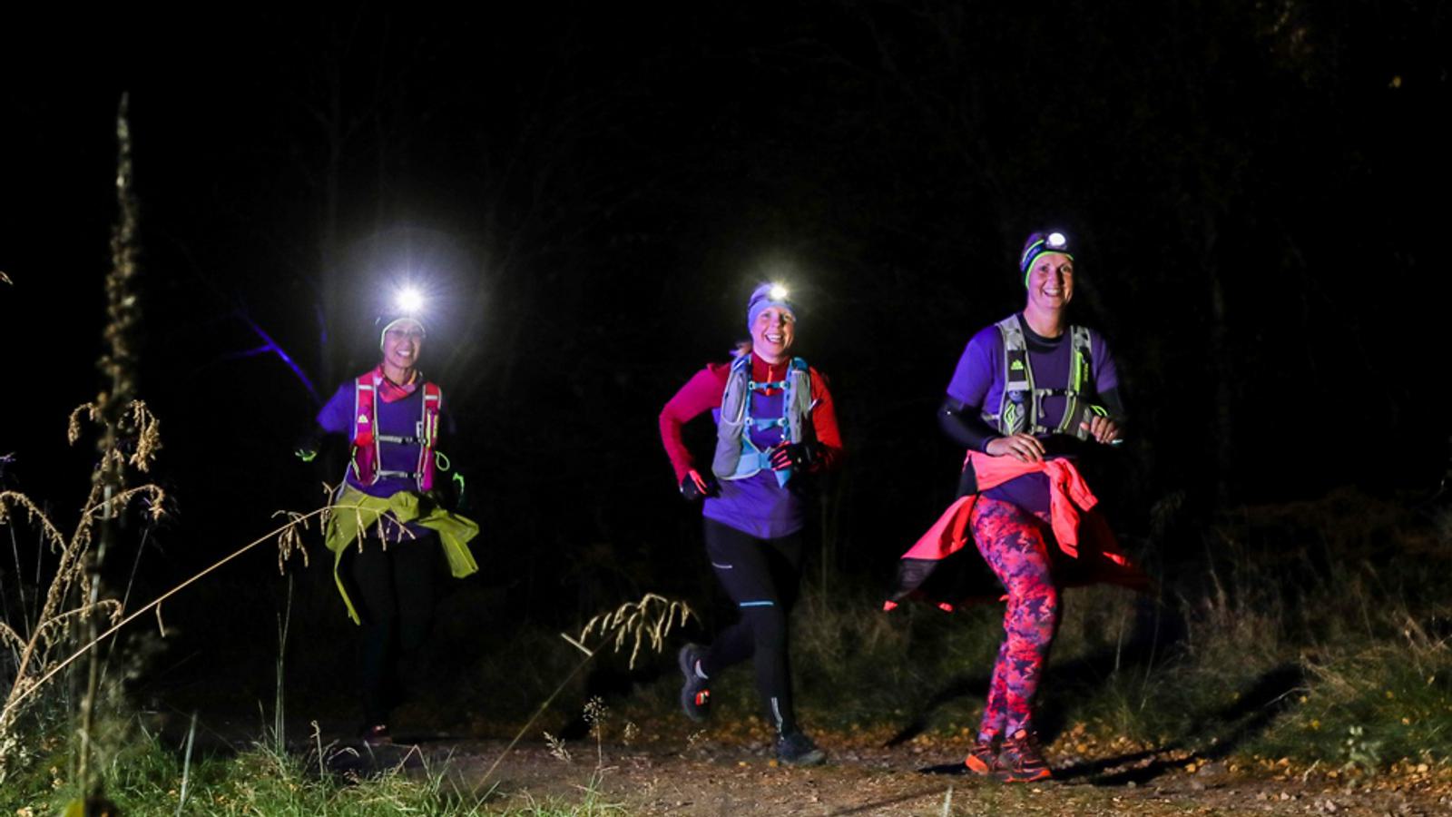 Three people are running on a trail at night. They are wearing headlamps and colorful running gear, with the path and surroundings dimly lit. The runners appear focused and energetic, with light vegetation visible around the trail.