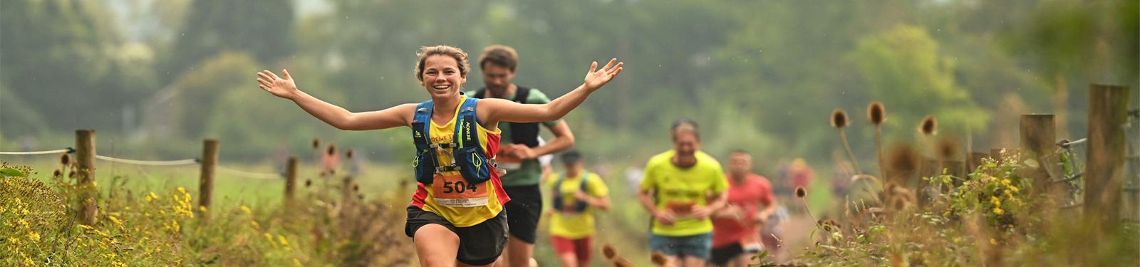 A woman in athletic gear runs joyfully on a dirt trail with outstretched arms, surrounded by other runners in a lush, green outdoor setting. Wildflowers and grass line the path, and the sky is overcast.