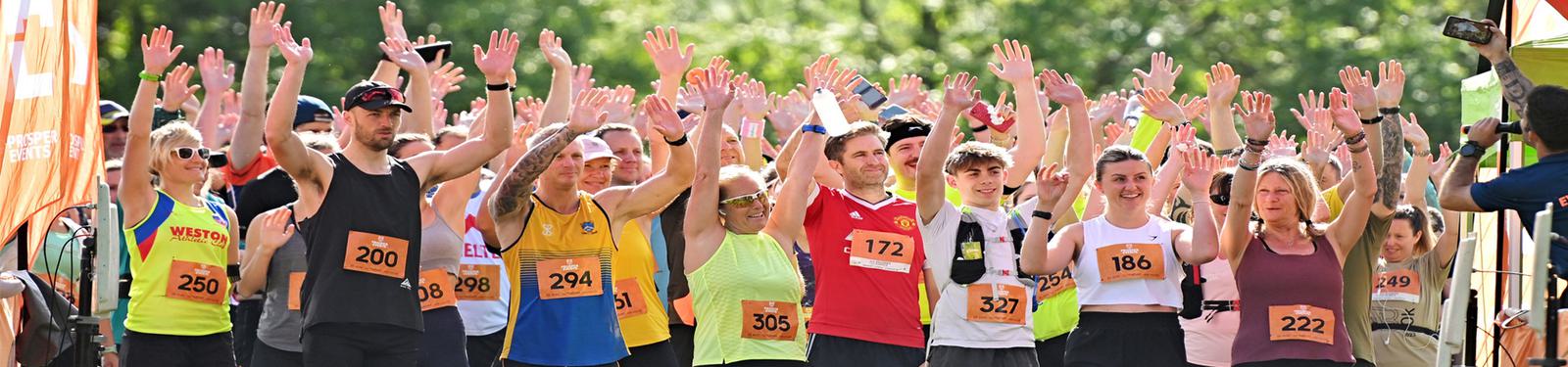 A group of marathon runners of various ages in colorful athletic gear raise their hands at the starting line. They are outdoors with green trees in the background, wearing race bibs.