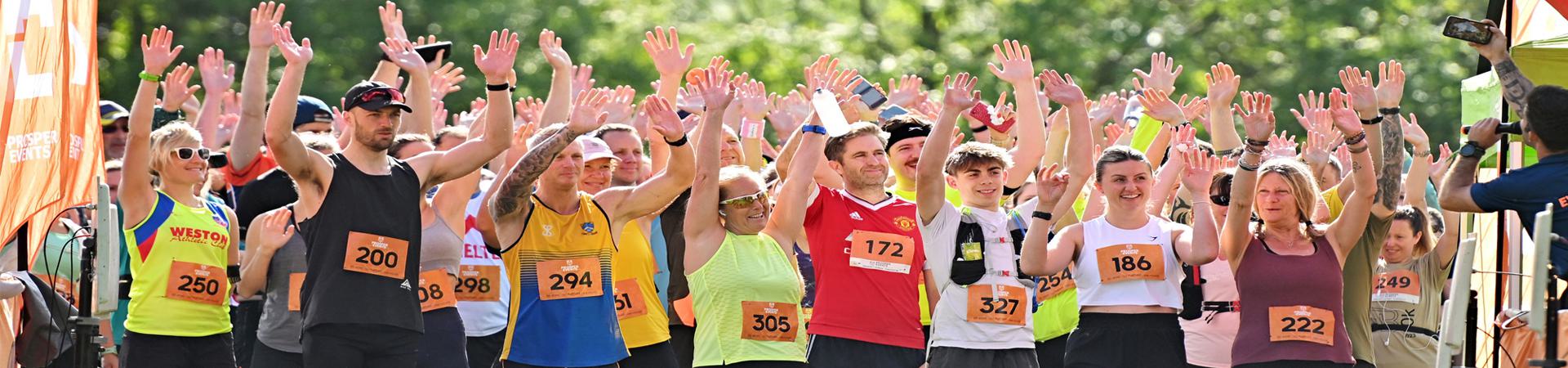 A group of marathon runners of various ages in colorful athletic gear raise their hands at the starting line. They are outdoors with green trees in the background, wearing race bibs.