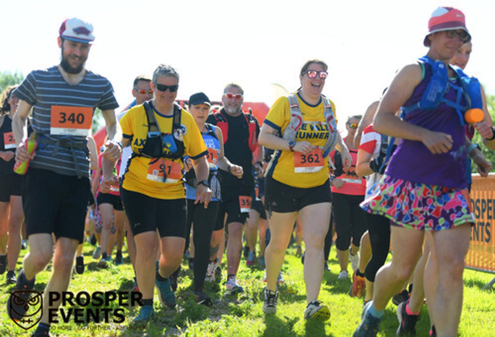 A group of runners participates in an outdoor race under sunny conditions. They wear various sports attire and numbers, and some carry hydration packs and water bottles. Event barriers are visible in the background along with the logo "Prosper Events.