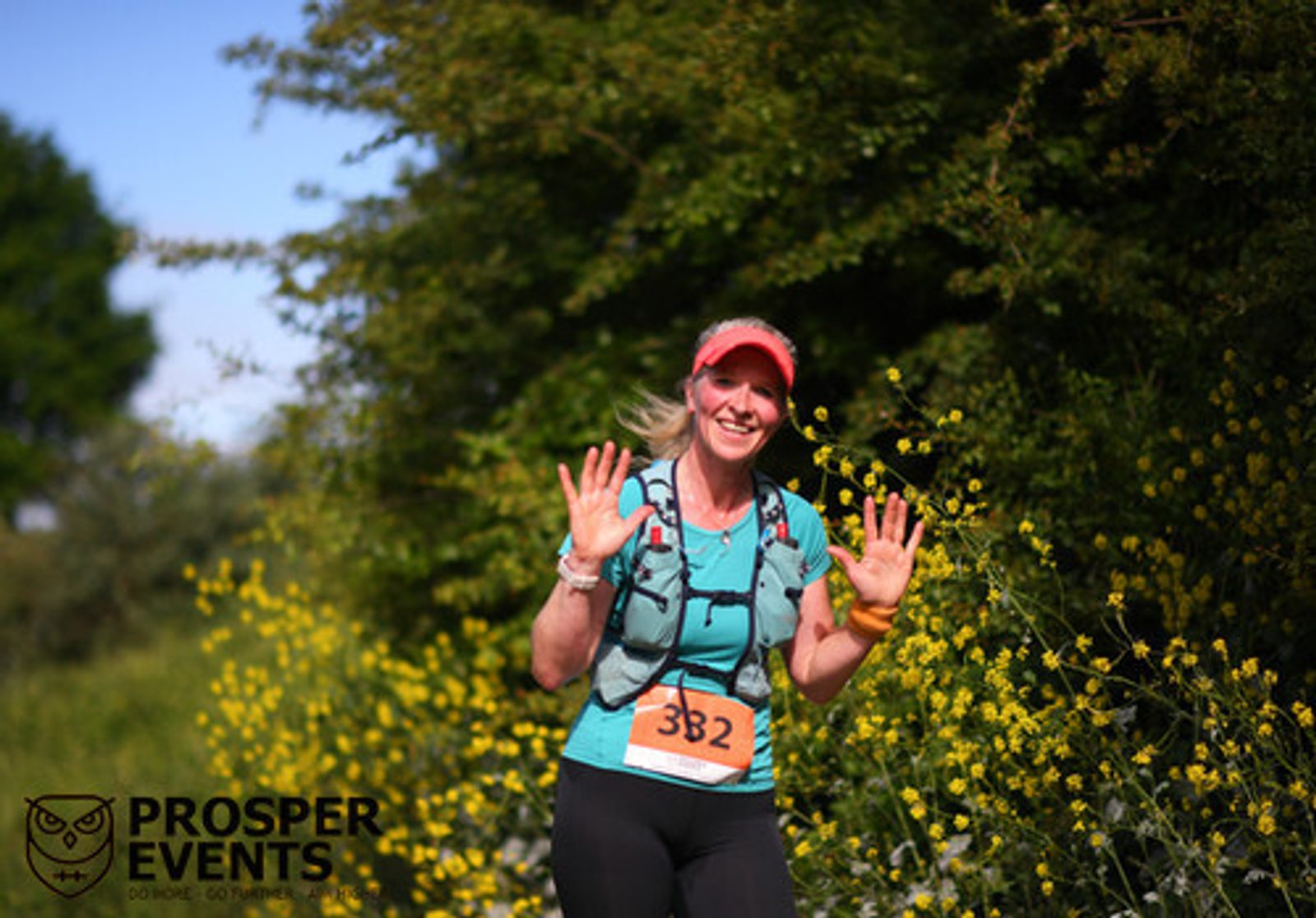 A smiling woman wearing a red cap, blue shirt, and race bib number 382 is running along a trail surrounded by yellow wildflowers. She is holding her hands up and appears to be waving. The logo "PROSPER EVENTS" is visible in the bottom left corner of the image.