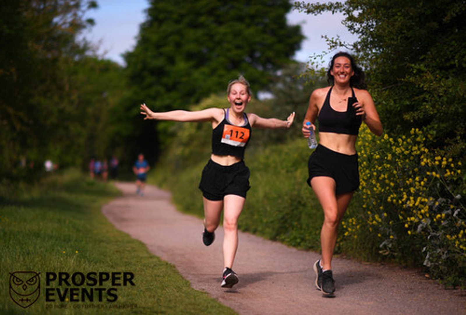 Two women in athletic wear are running along a tree-lined path, both smiling and appearing joyful. One woman, wearing race bib number 112, has her arms outstretched. The other woman holds a water bottle. In the background, more runners can be seen. The logo "PROSPER EVENTS" is in the corner.
