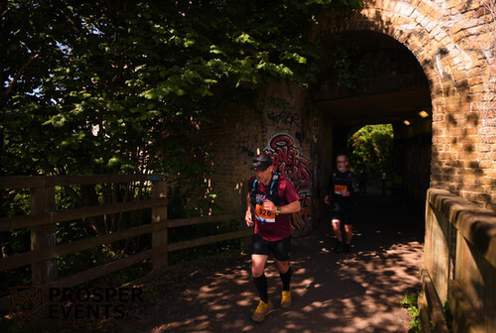 Runners jog through a tunnel with graffiti under a brick bridge during a sunny outdoor race. Trees and foliage surround the path, casting shadows. A person in the foreground wears a red shirt and a blue backpack, followed closely by others.