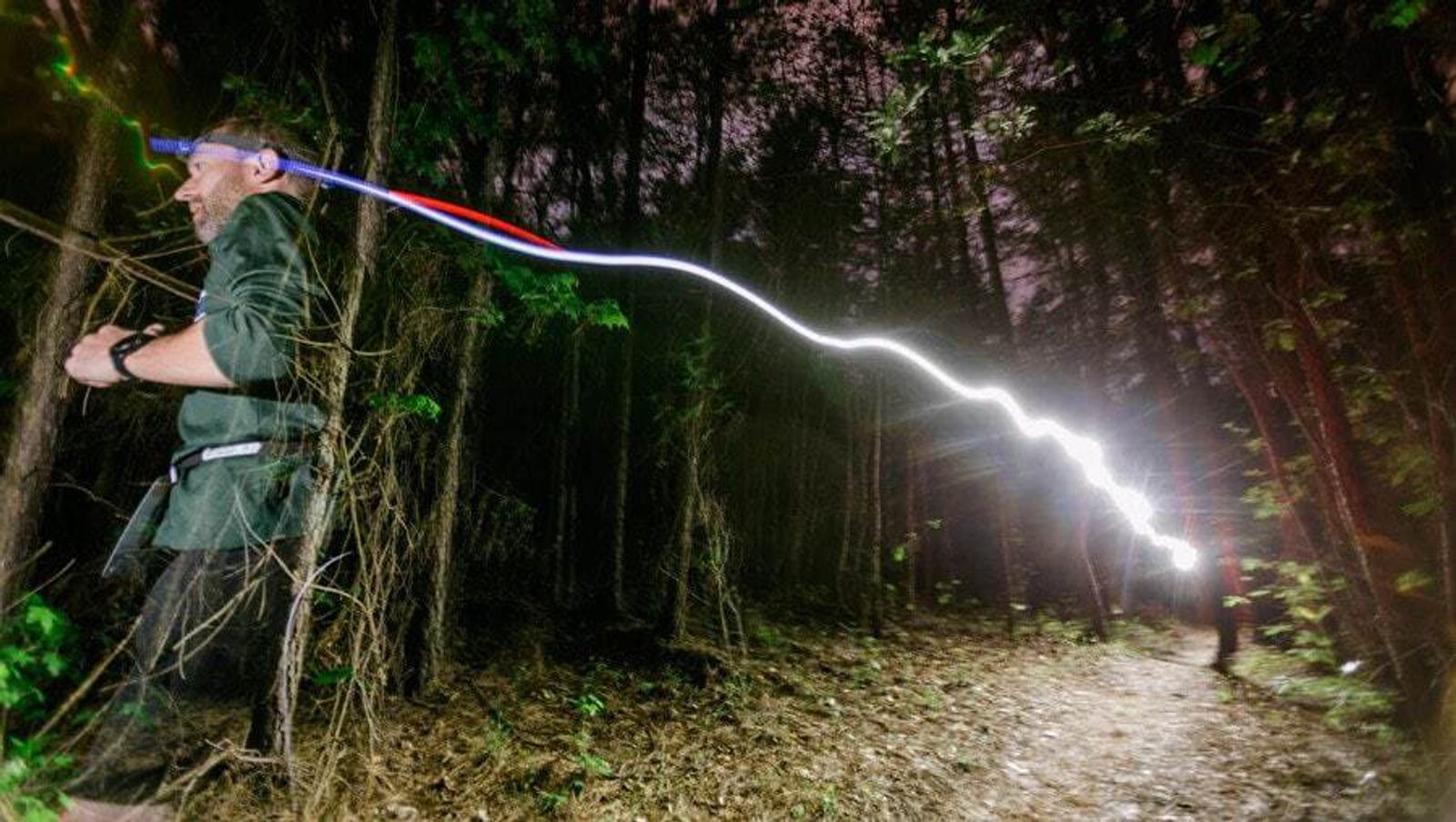 A person runs through a dense, dark forest at night, wearing a headlamp that leaves a bright, winding trail of light behind them. The forest floor is covered with leaves and foliage, and the sky is dim with very little visible through the trees.