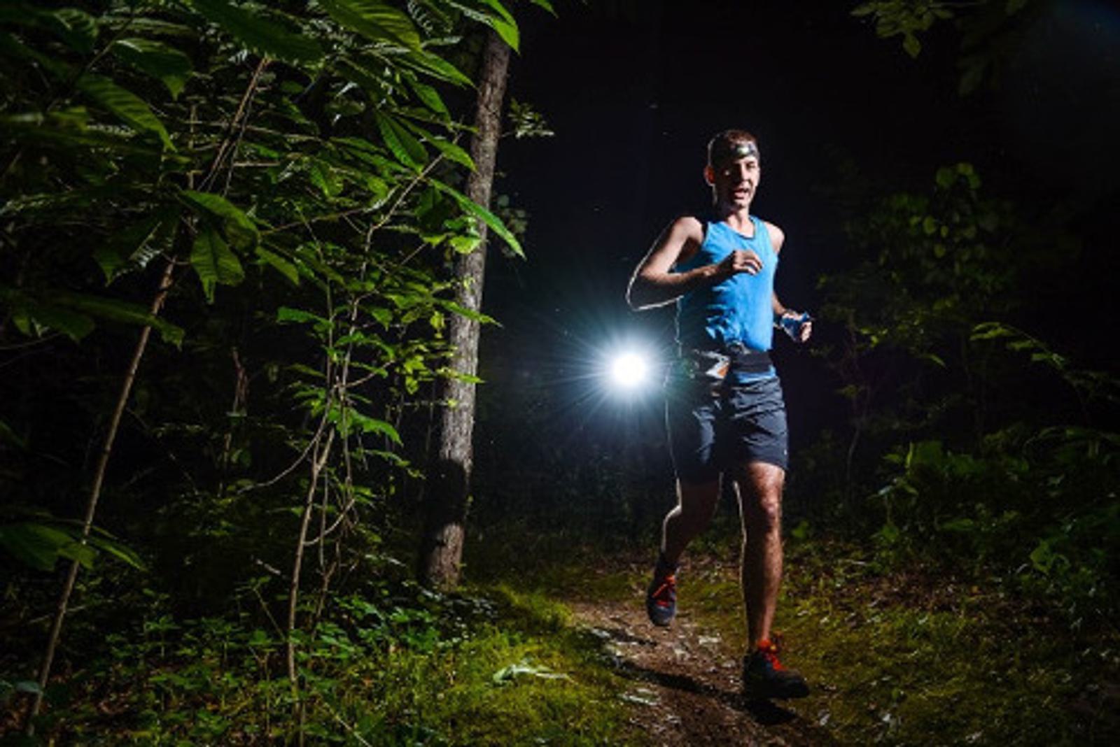 A man in a blue tank top and black shorts is running on a forest trail at night. He is illuminated by a headlamp, with dense green foliage surrounding him and a bright light shining from behind. The scene captures a sense of determination and adventure.