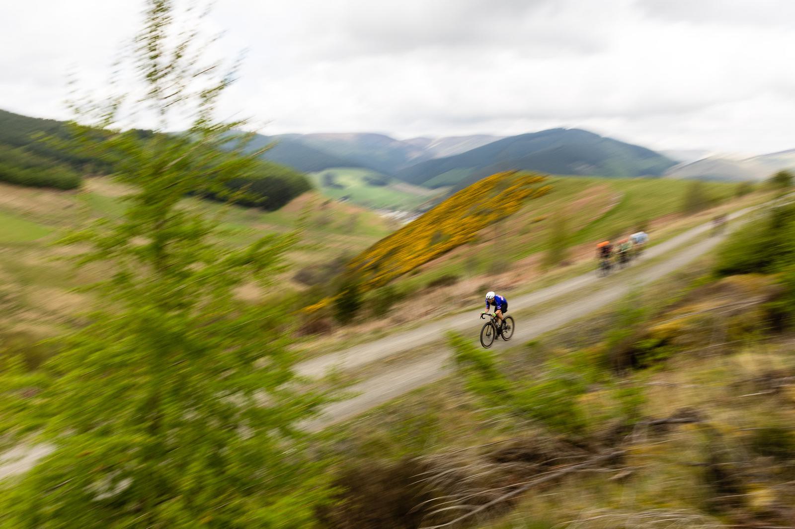 A cyclist in blue races along a winding road through a mountainous landscape. The motion blur emphasizes speed. The scene is surrounded by lush greenery and distant rolling hills under a cloudy sky. More cyclists follow in the background.