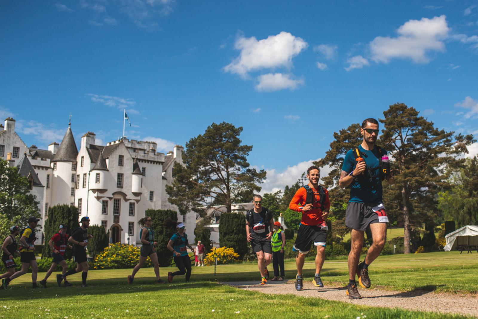 Runners participating in a race pass by a historic building with turrets, set against a clear blue sky. Lush greenery surrounds the path, with trees and flowers in the background.