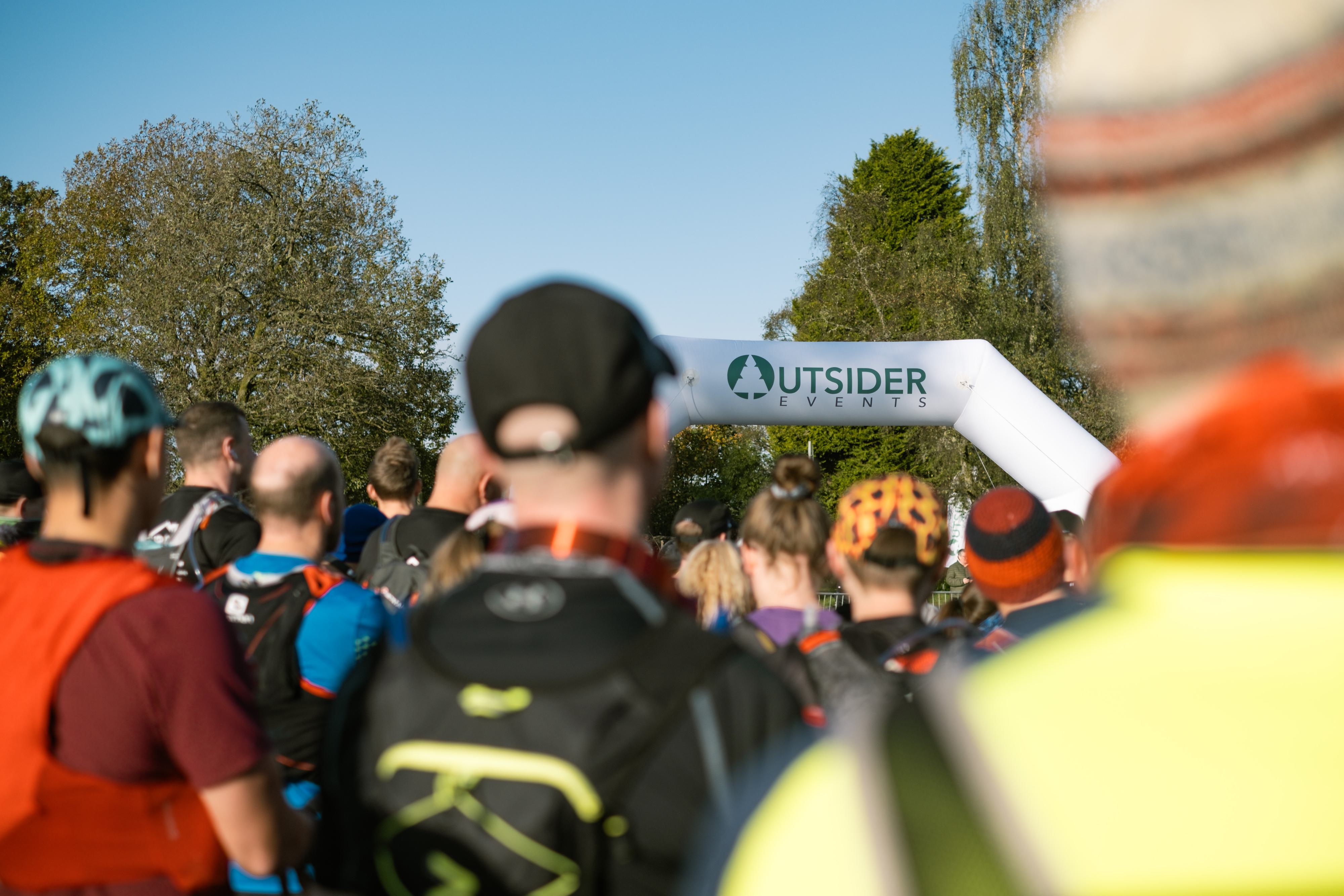Participants gather at the start line of an outdoor event hosted by Outsider Events. The crowd, wearing colorful athletic gear, stands in front of a large inflatable arch. Trees and a clear blue sky are in the background.