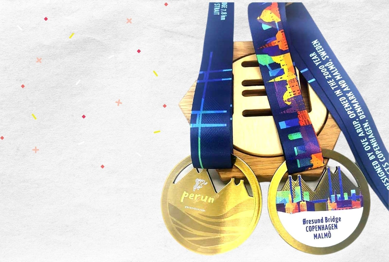 Two medals hanging side by side on blue ribbons against a white background. The left medal is gold with a minimalist runner icon and "Perun." The right medal features a colorful image of a bridge with text referencing the Øresund Bridge between Copenhagen and Malmö.