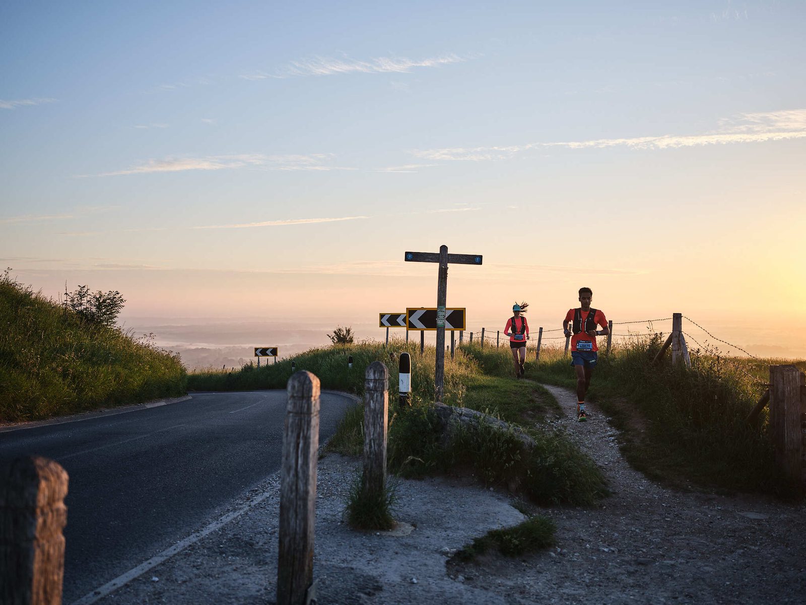 Two runners are on a sunlit trail next to a road with wooden posts and a signpost in the background. The sky is clear with a warm, glowing sunset. The scene is serene and captures the runners mid-action, immersed in the natural, open surroundings.
