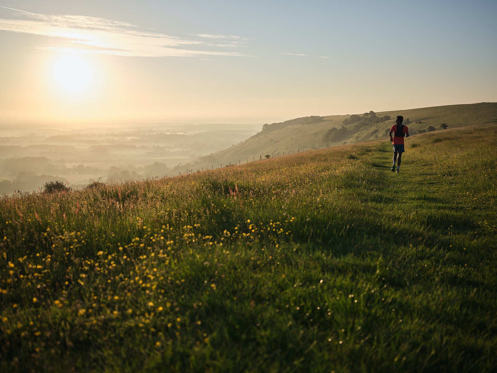 A person jogs along a narrow grassy path on a hillside during sunrise. The sky is clear with a few wispy clouds, and the landscape is dotted with wildflowers. The sun casts a warm, golden light over the rolling hills and distant horizon.