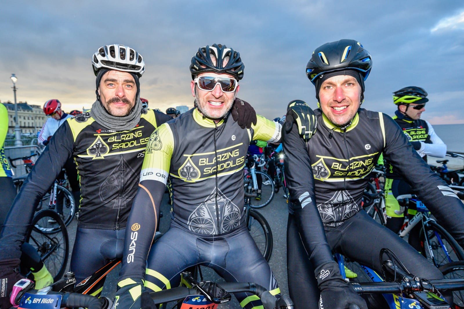 Three cyclists wearing matching "Blazing Saddles" cycling jerseys and helmets smile and pose together with their bikes. They are part of a large group of cyclists gathered on a road, with a cloudy sky in the background.