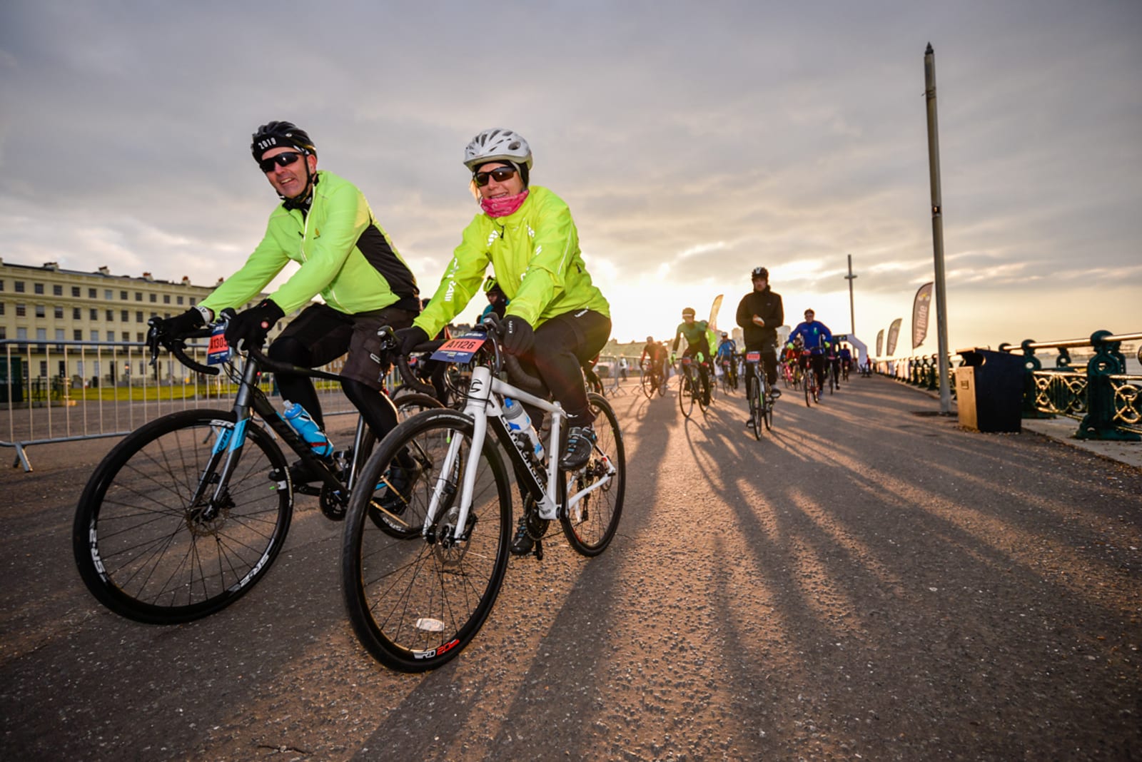 Cyclists in bright yellow jackets ride along a paved boardwalk at sunrise. The sky is partly cloudy and they are among other cyclists in the background. A large building is visible on the left, and shadows stretch across the ground.