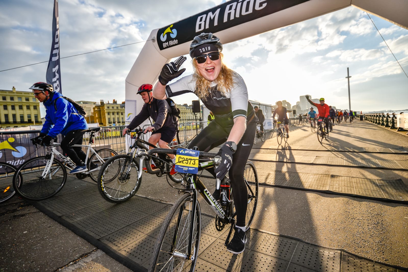A cyclist wearing black and gray attire waves enthusiastically while crossing under an arch marked "BM Ride." Other cyclists ride beside and behind them on a scenic boardwalk by the seaside. The event appears lively and well-attended, with clear, sunny weather.