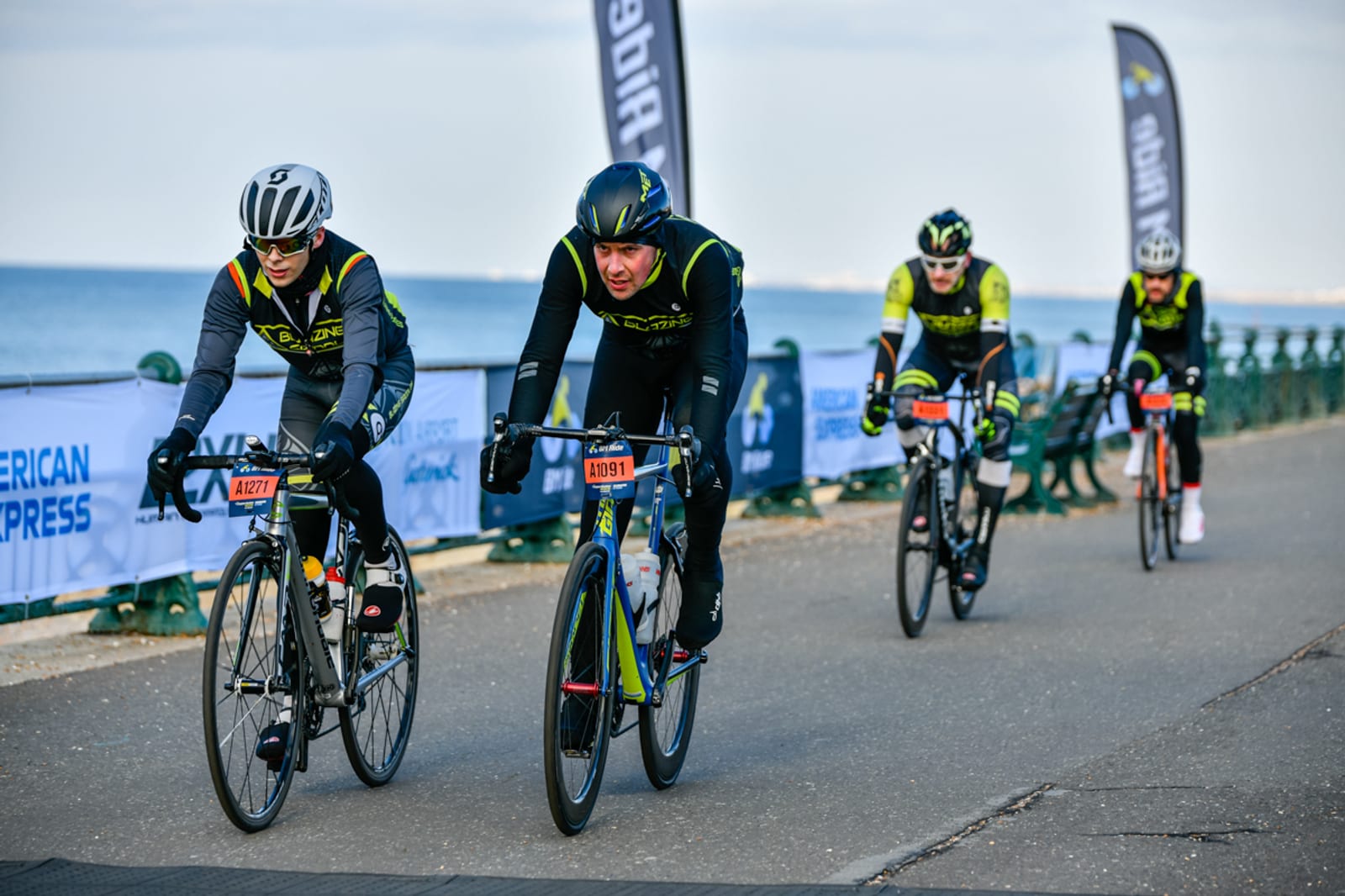 A group of cyclists wearing racing gear competes in a triathlon along a waterfront path. Banners and railings line the path, and the water is visible in the background. The riders are focused and pushing forward on their bicycles.