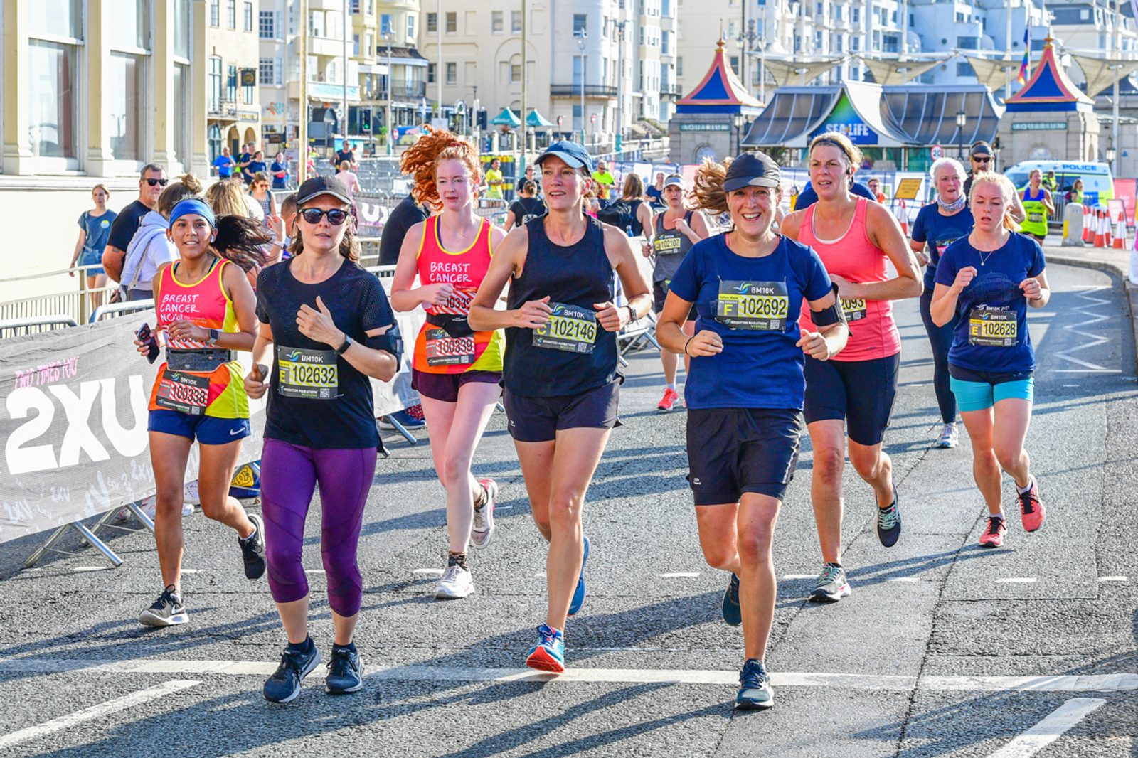 A group of runners, both men and women, participating in a race on a sunny day. They appear joyful and energetic, some smiling towards the camera. The background shows city buildings, spectators, and race barriers along the street.