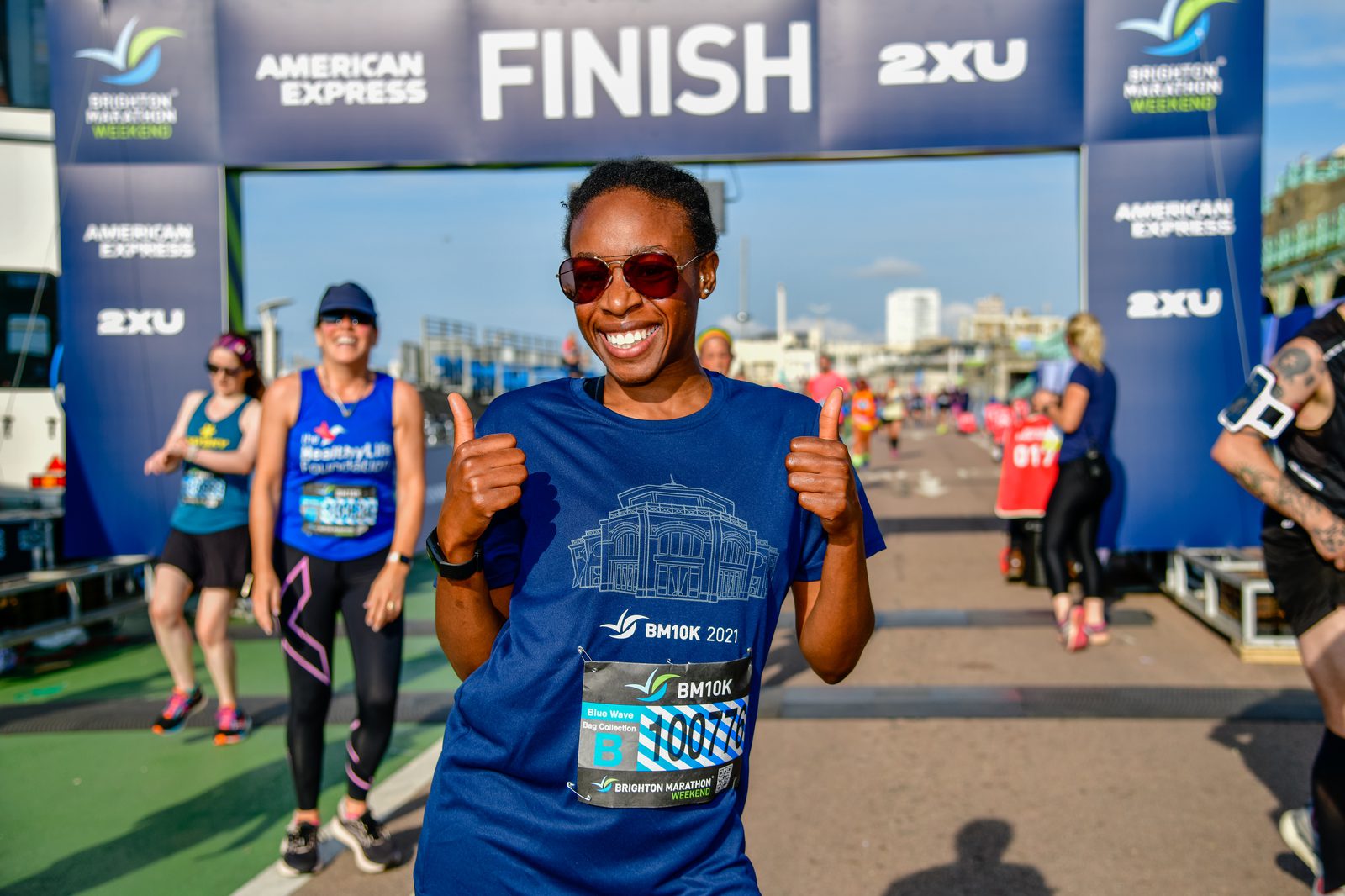 A smiling runner wearing sunglasses and a blue shirt stands near the finish line of a marathon, holding two thumbs up. The finish line banner and background include text for sponsors and the event, with other runners and spectators visible.
