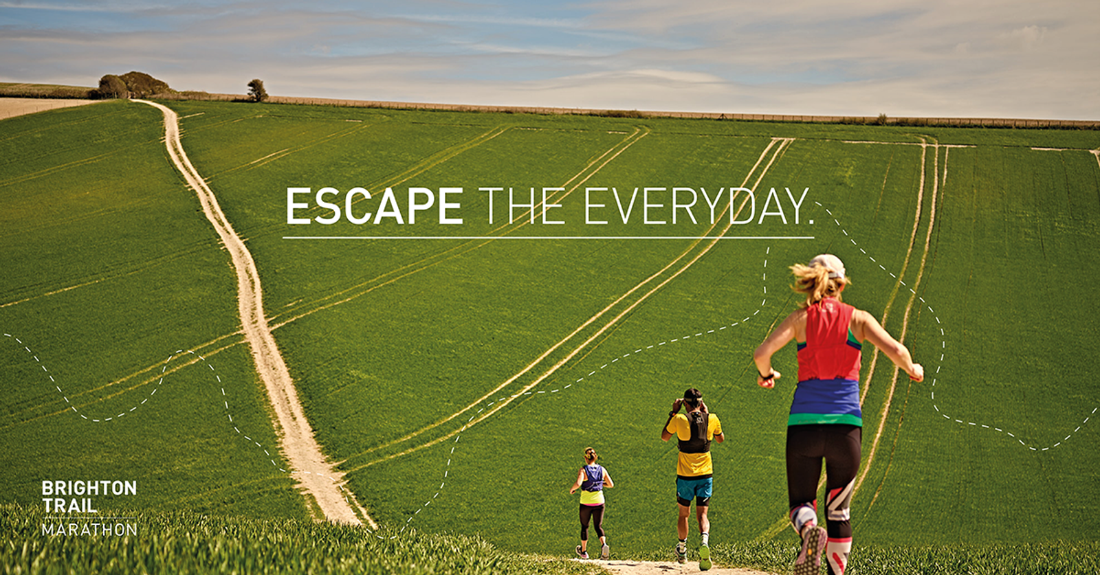Three runners are traversing a sunlit, green countryside trail with expansive fields around them. The text "ESCAPE THE EVERYDAY" is prominently displayed in the sky above. A logo in the bottom left corner reads "BRIGHTON TRAIL MARATHON.