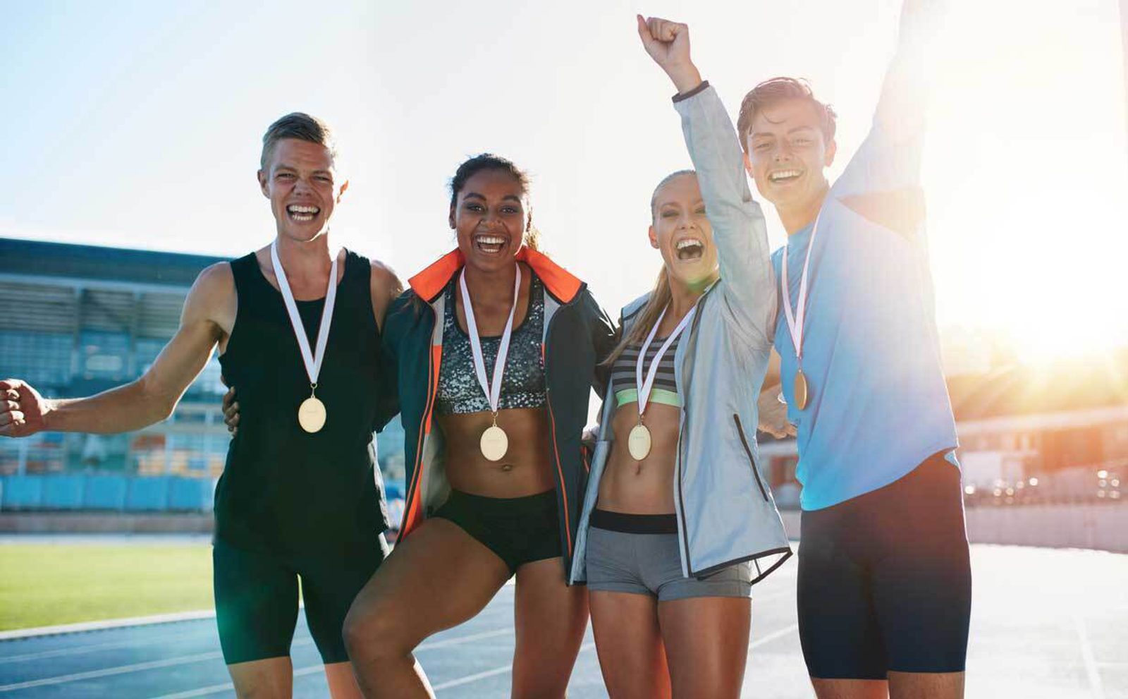 Four athletes standing together on a sports track, smiling and cheering. They are wearing athletic gear and each has a gold medal around their neck. Sunlight shines brightly in the background.