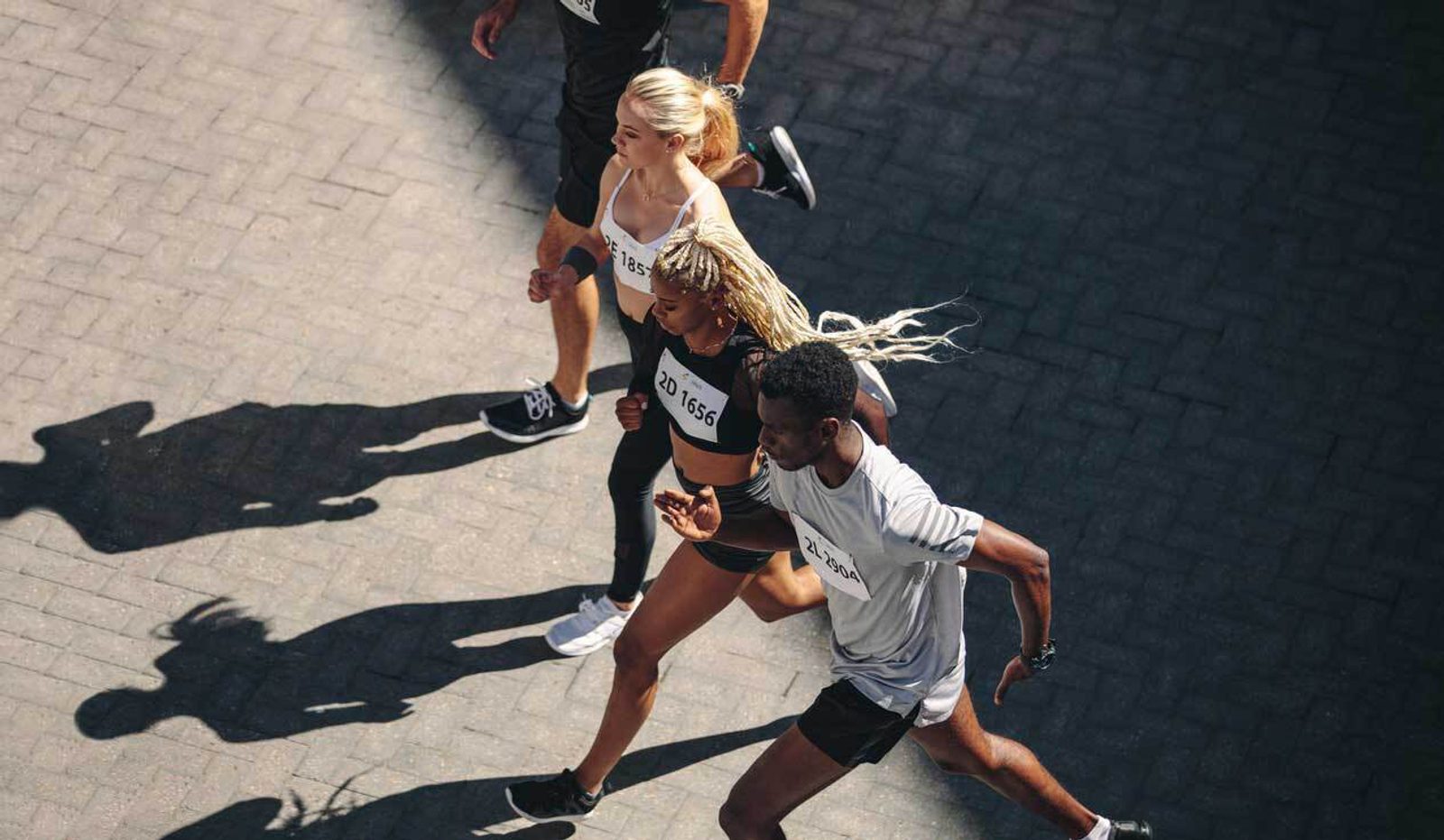 Top view of three runners in sportswear participating in a race. They are running closely together on a stone-paved path, with race numbers visible on their shirts. Their shadows stretch out on the ground in the sunlight.