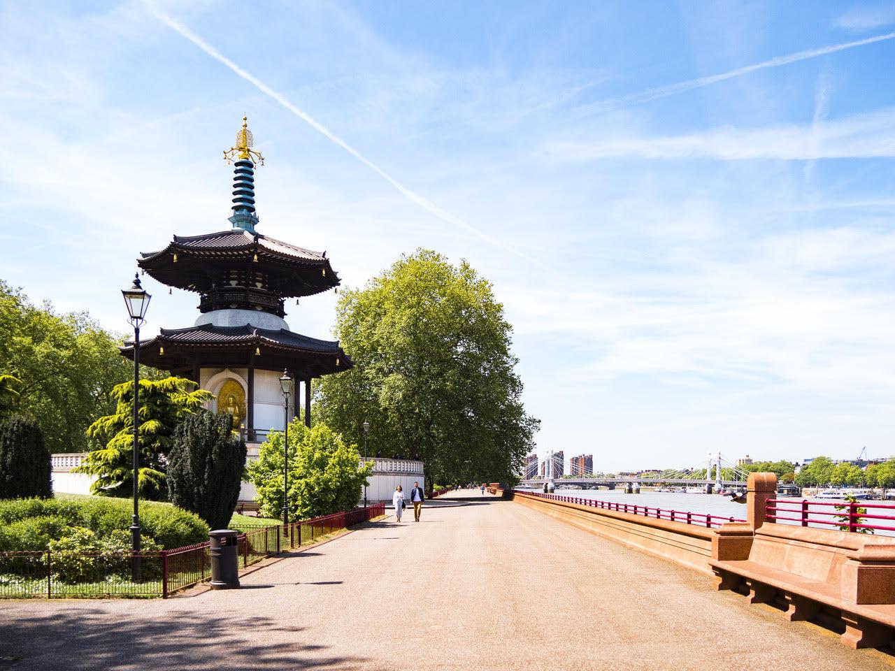 A serene park scene with a traditional Japanese pagoda on the left, surrounded by lush green trees. A wide pathway leads into the distance along a river with a blue sky overhead. A few people are strolling, and a red railing runs alongside the river.