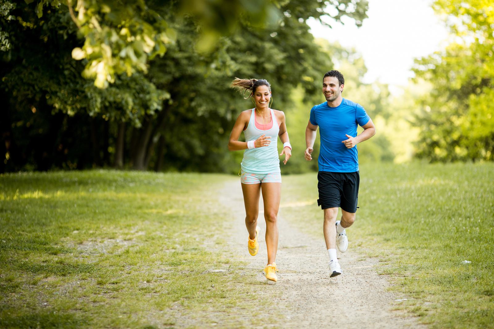 A woman and a man are jogging together on a dirt path in a lush, green park. The woman, wearing a light blue tank top and shorts, and the man, in a blue t-shirt and black shorts, are smiling and appear to be enjoying the exercise on a sunny day.