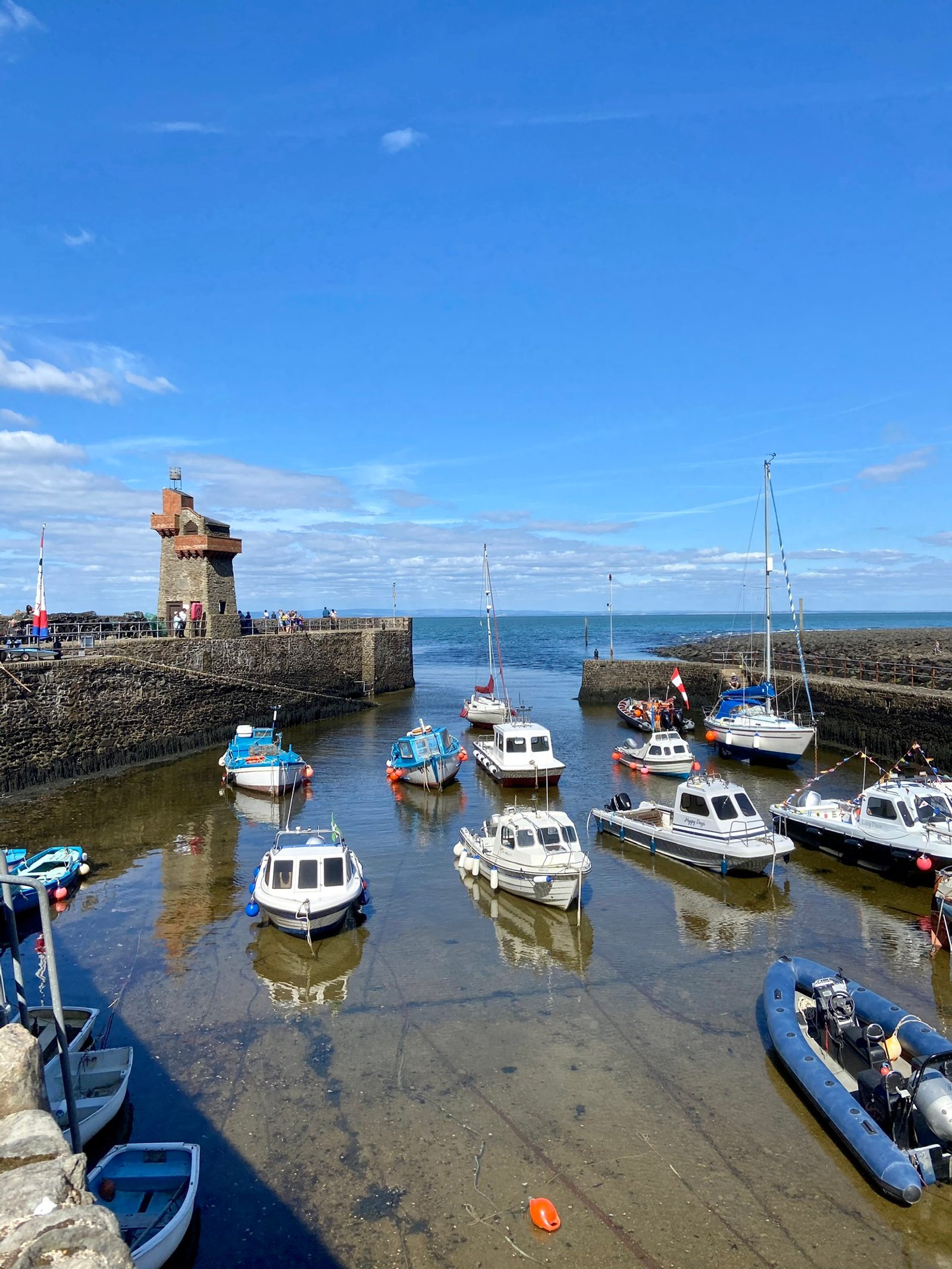 A small harbor filled with various boats, including yachts and motorboats, tied to docks or anchored in shallow, calm waters. A stone tower stands at the entrance to the harbor under a partly cloudy blue sky.