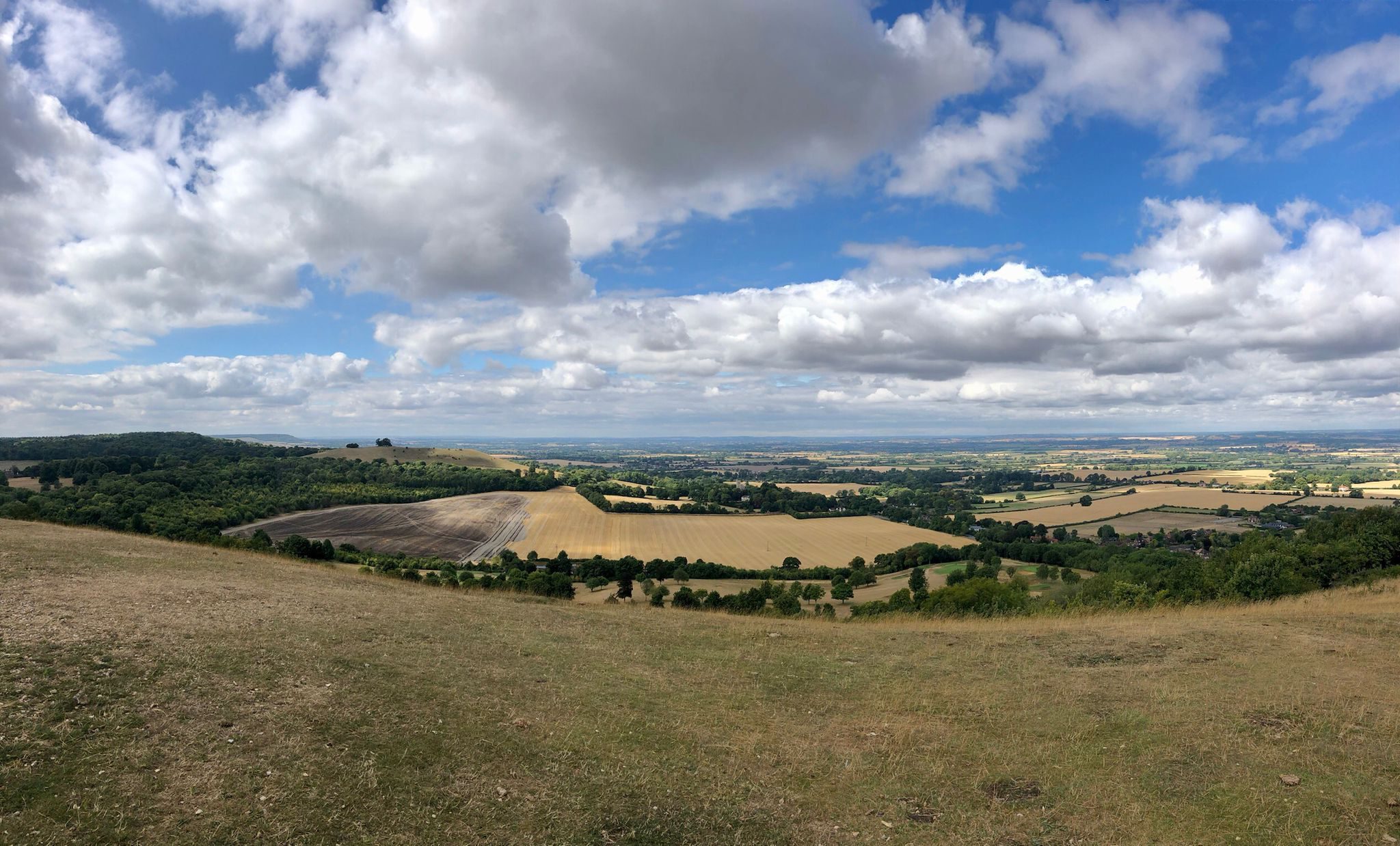A scenic view of a vast countryside landscape under a partly cloudy sky. Rolling hills covered with fields of varying shades of green and brown stretch into the distance, with clusters of trees dotting the terrain. The horizon shows more hills and hints of distant villages.