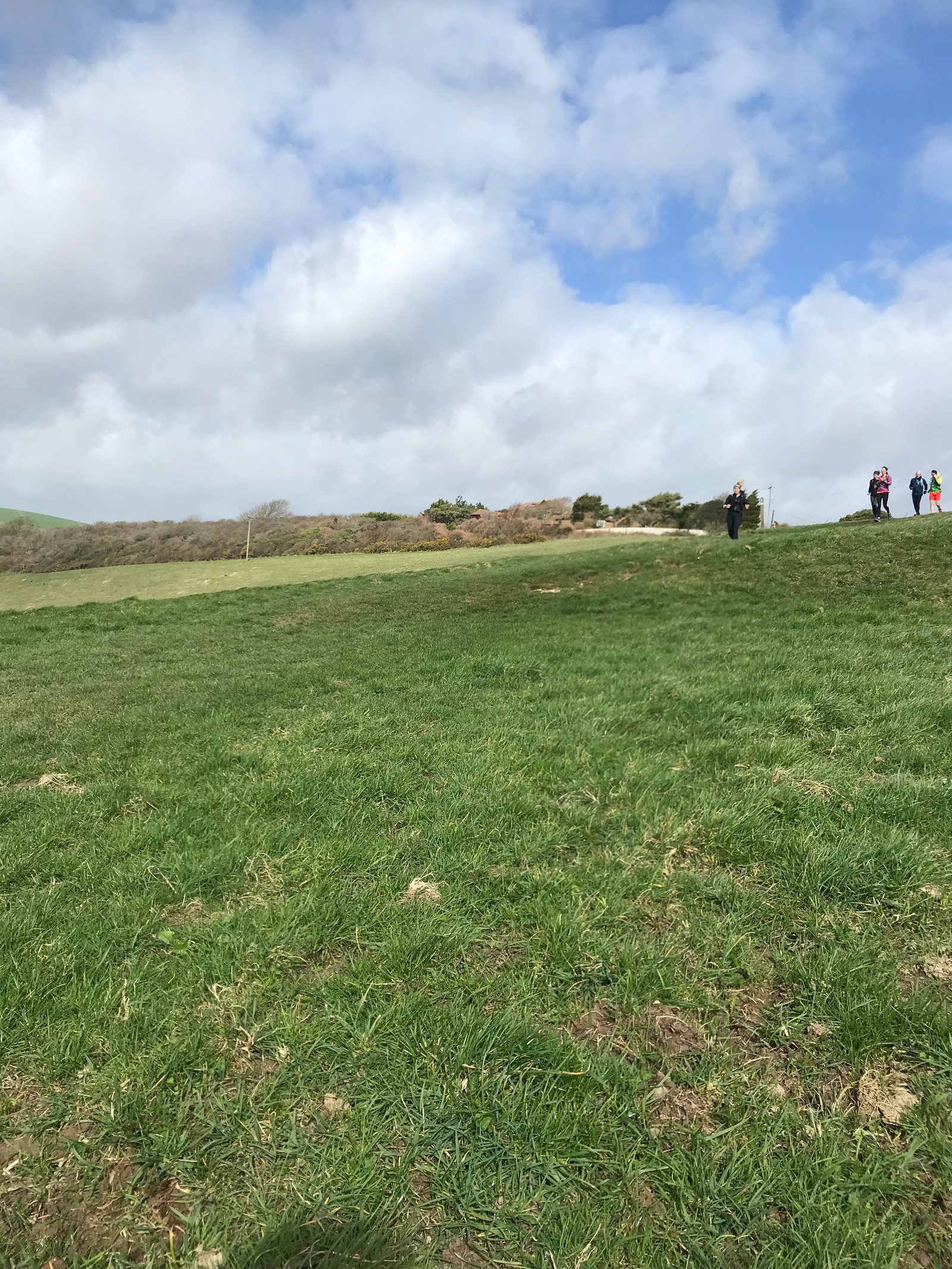A vast green field with patches of grass and a gentle slope leading up to a ridgeline. A group of people are walking along the hilltop trail under a partly cloudy sky, enjoying a hike on a breezy day. Bushes and small trees are scattered in the background.