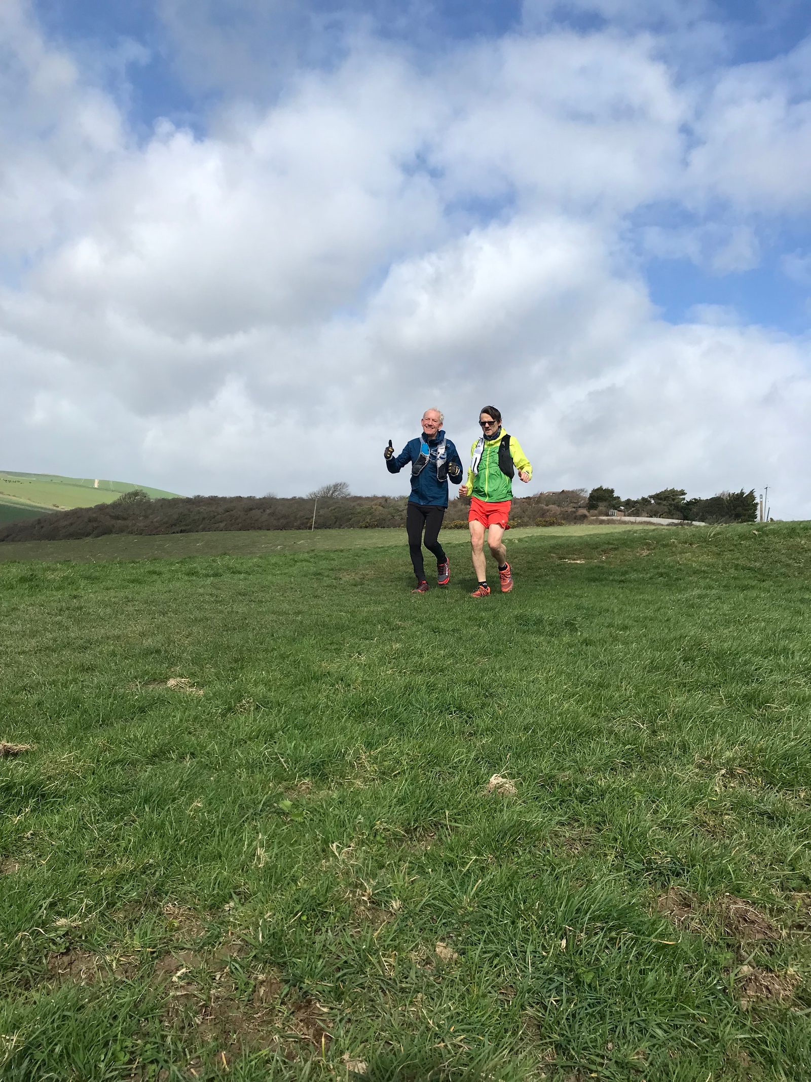 Two people are running on a grassy field under a partly cloudy sky. One is wearing a blue jacket, black leggings, and a gray beanie, while the other is in a neon jacket, red shorts, and sunglasses. They smile and appear to be enjoying their run.
