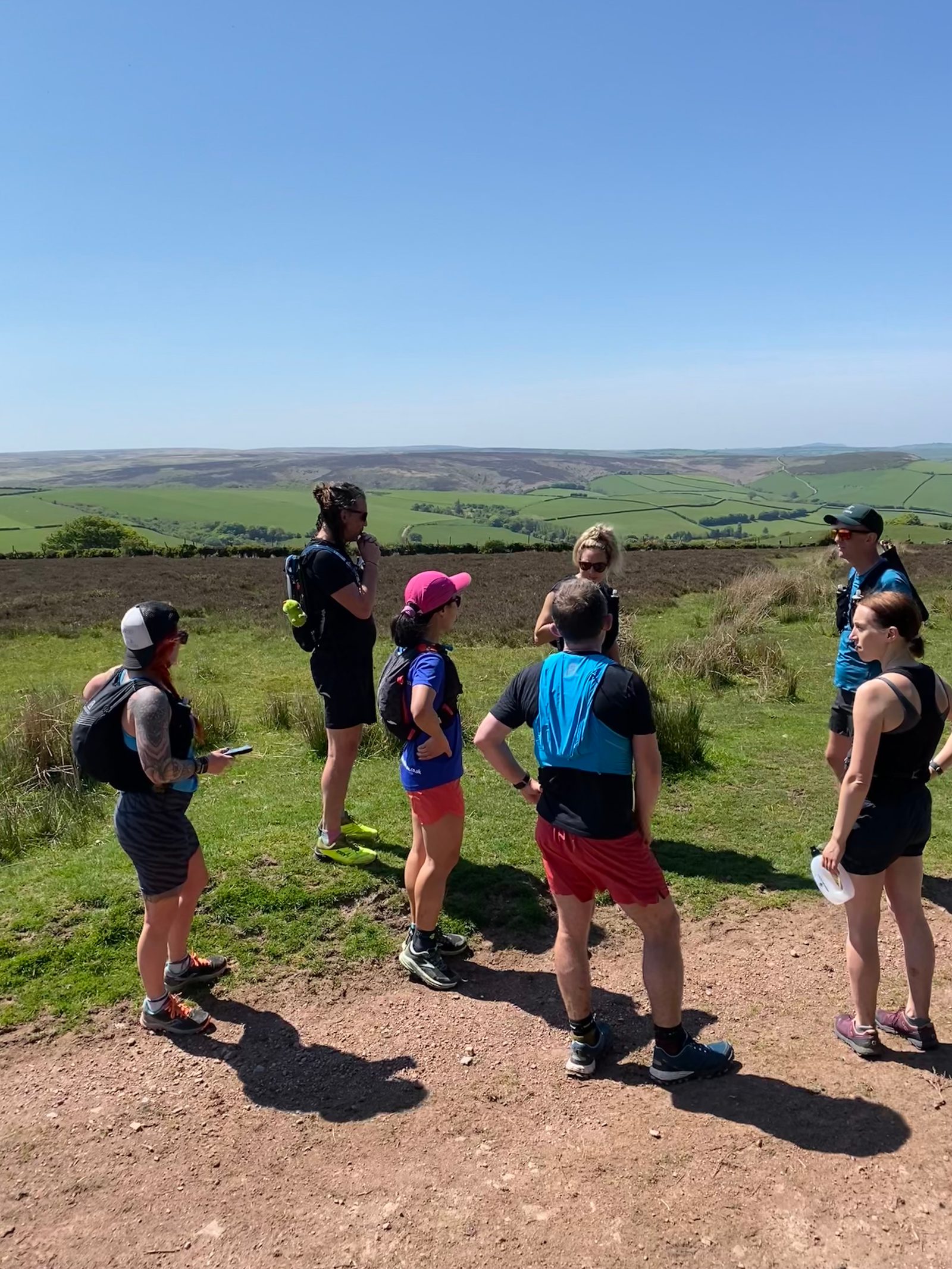 A group of eight people wearing athletic gear stand on a sunny trail in a green countryside landscape. Some have backpacks and are holding maps. They are gathered in a semi-circle, talking, with rolling hills and a clear blue sky in the background.