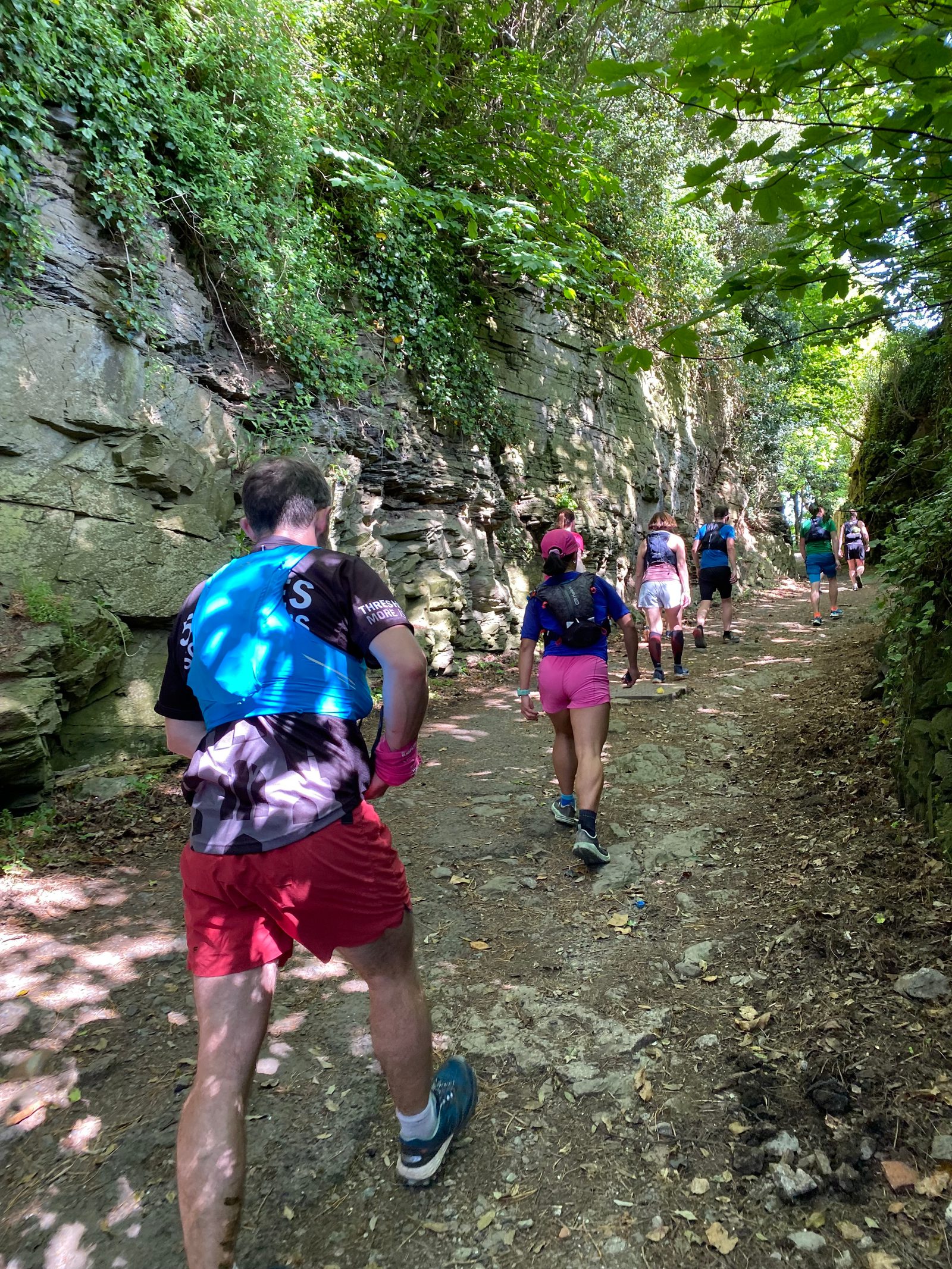 A group of people wearing athletic gear hike up a rocky and shaded forest trail. They are surrounded by dense greenery and large rock walls. The trail surface appears rough and natural. Sunlight filters through the leaves above.