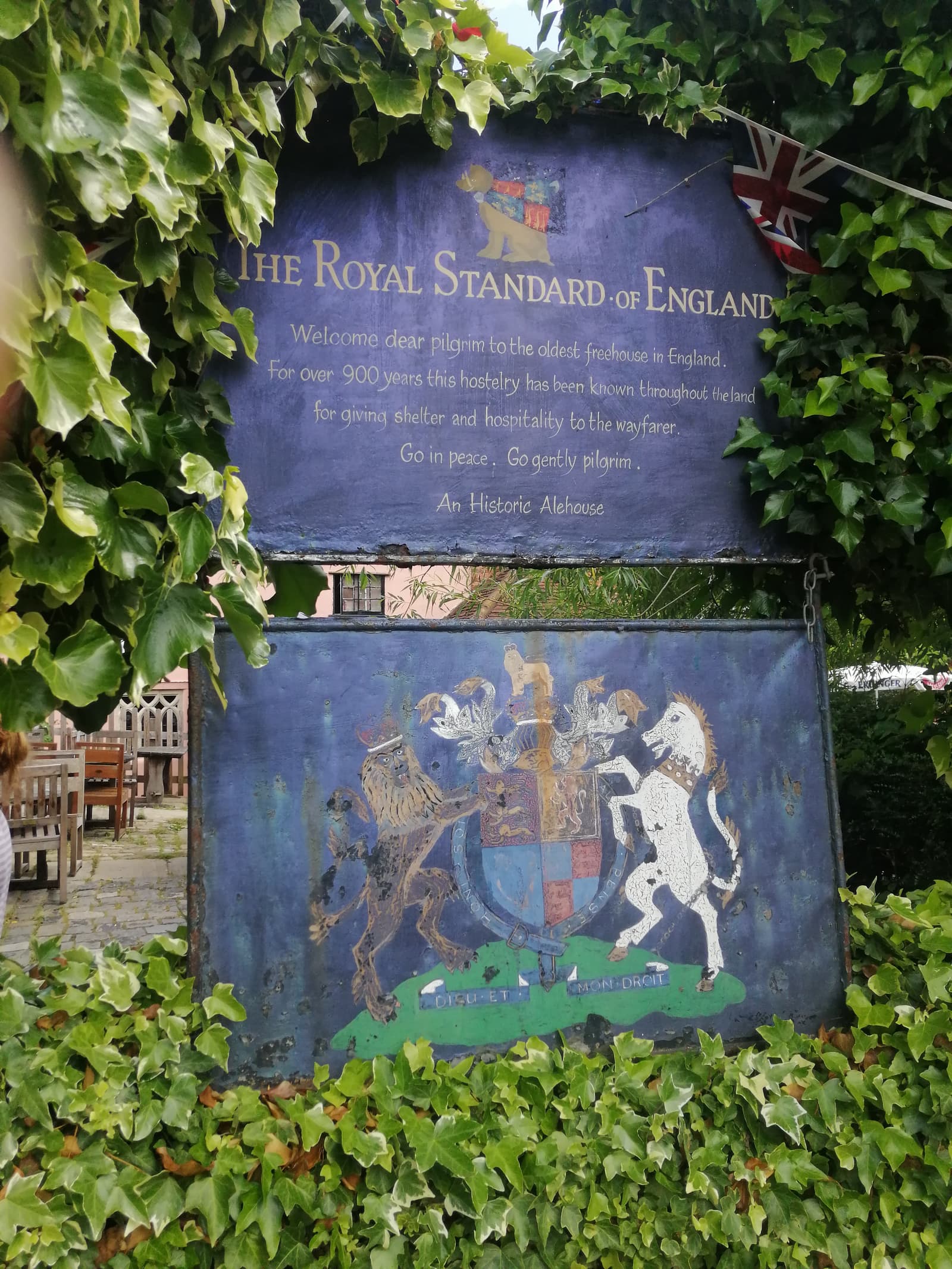 A weathered sign surrounded by ivy reads, "The Royal Standard of England. Welcome dear pilgrim to the oldest freehouse in England. For over 900 years this hostelry has been known throughout the land for giving shelter and hospitality to the wayfarer. Go in peace. Go gently pilgrim. An Historic Alehouse." Beneath the text is an illustrated crest featuring a lion and a unicorn. Nearby, wooden tables and benches are partially visible.