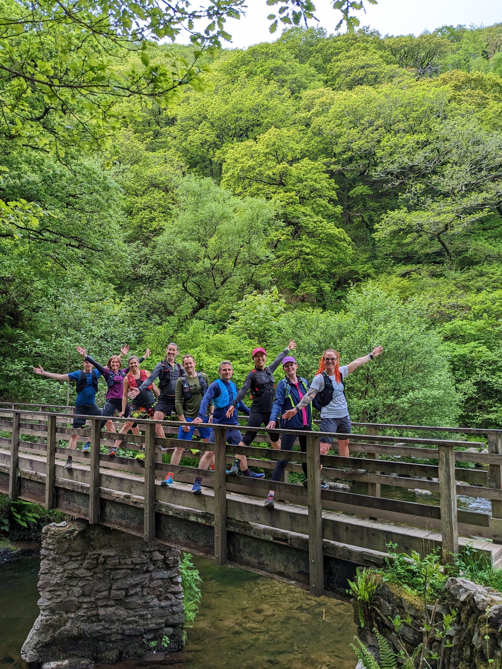 A group of ten people posing cheerfully on a wooden bridge surrounded by lush, green forest. Some members of the group are making playful gestures, and everyone appears to be enjoying the outdoor environment. The bridge crosses over a small stream.