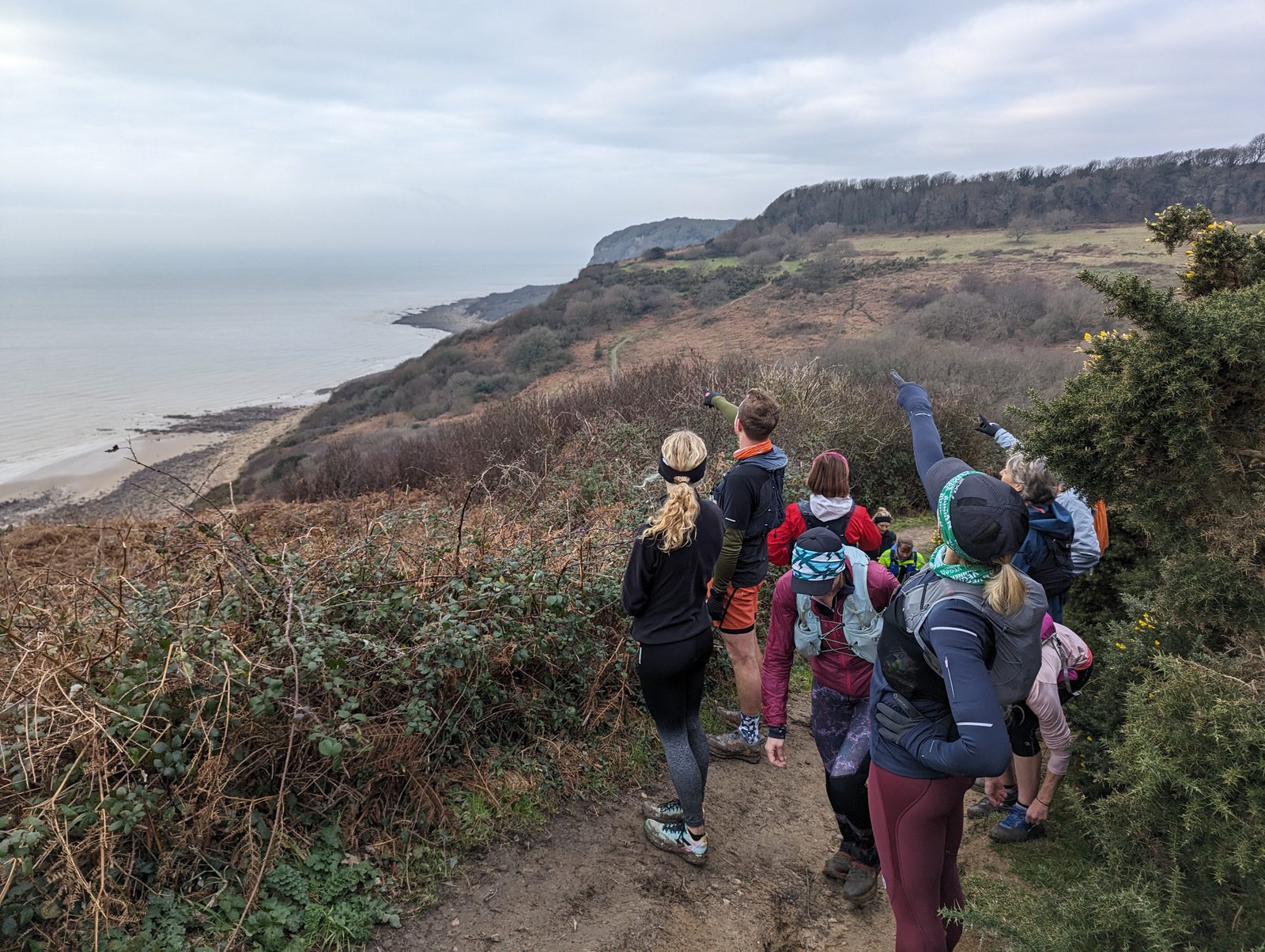 A group of people in athletic attire stand on a dirt trail near a coastal cliff, overlooking the sea. They are pointing towards the horizon, with lush greenery and a hilly landscape in the background under a cloudy sky.