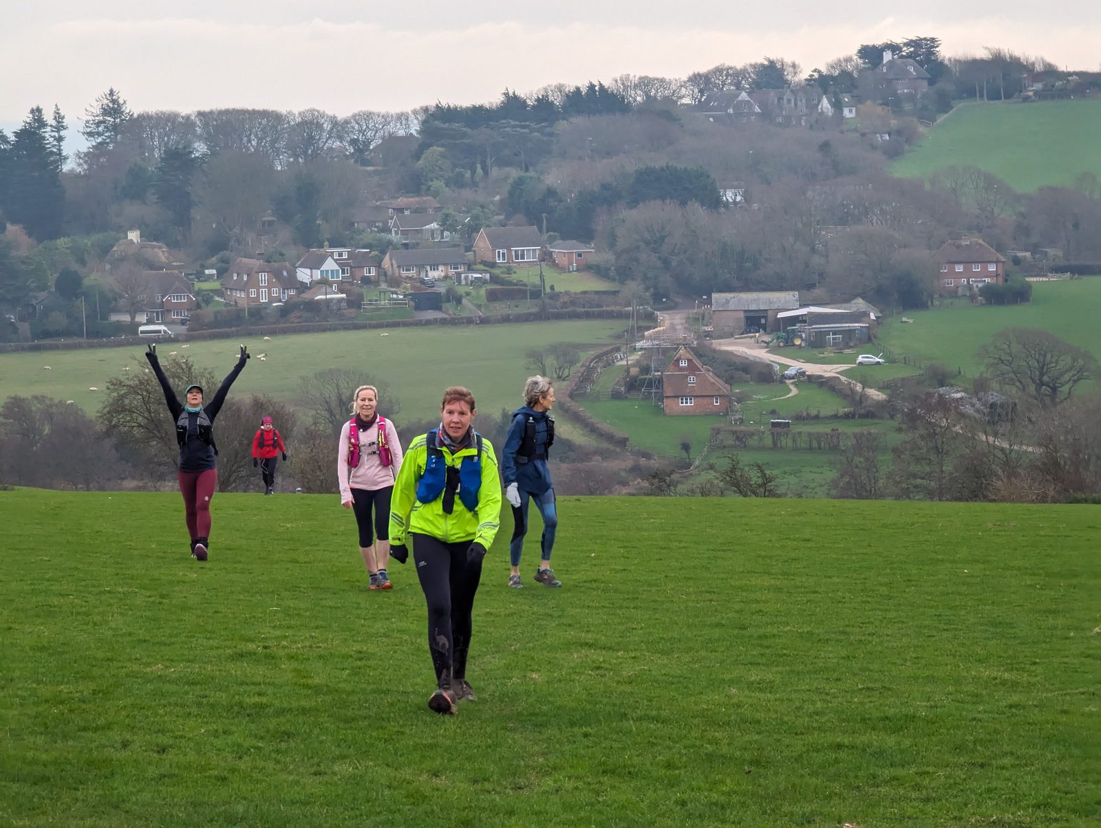 A group of hikers, dressed in outdoor gear, walk across a grassy field. They approach the camera with smiles and raised arms. In the background, a rural landscape features houses, trees, and rolling hills under a cloudy sky.