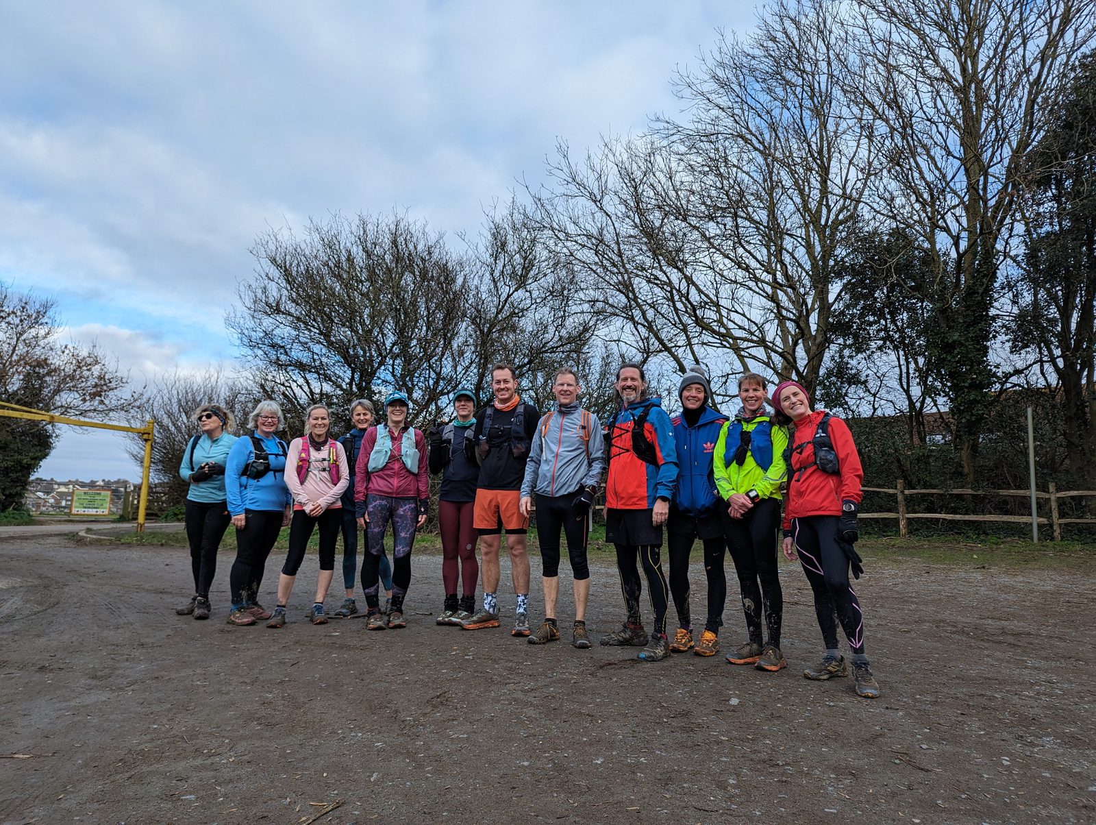A group of twelve people dressed in outdoor athletic gear stand in a row on a dirt path, smiling at the camera. They appear to be ready for a hike or run. Bare trees and a cloudy sky form the background.