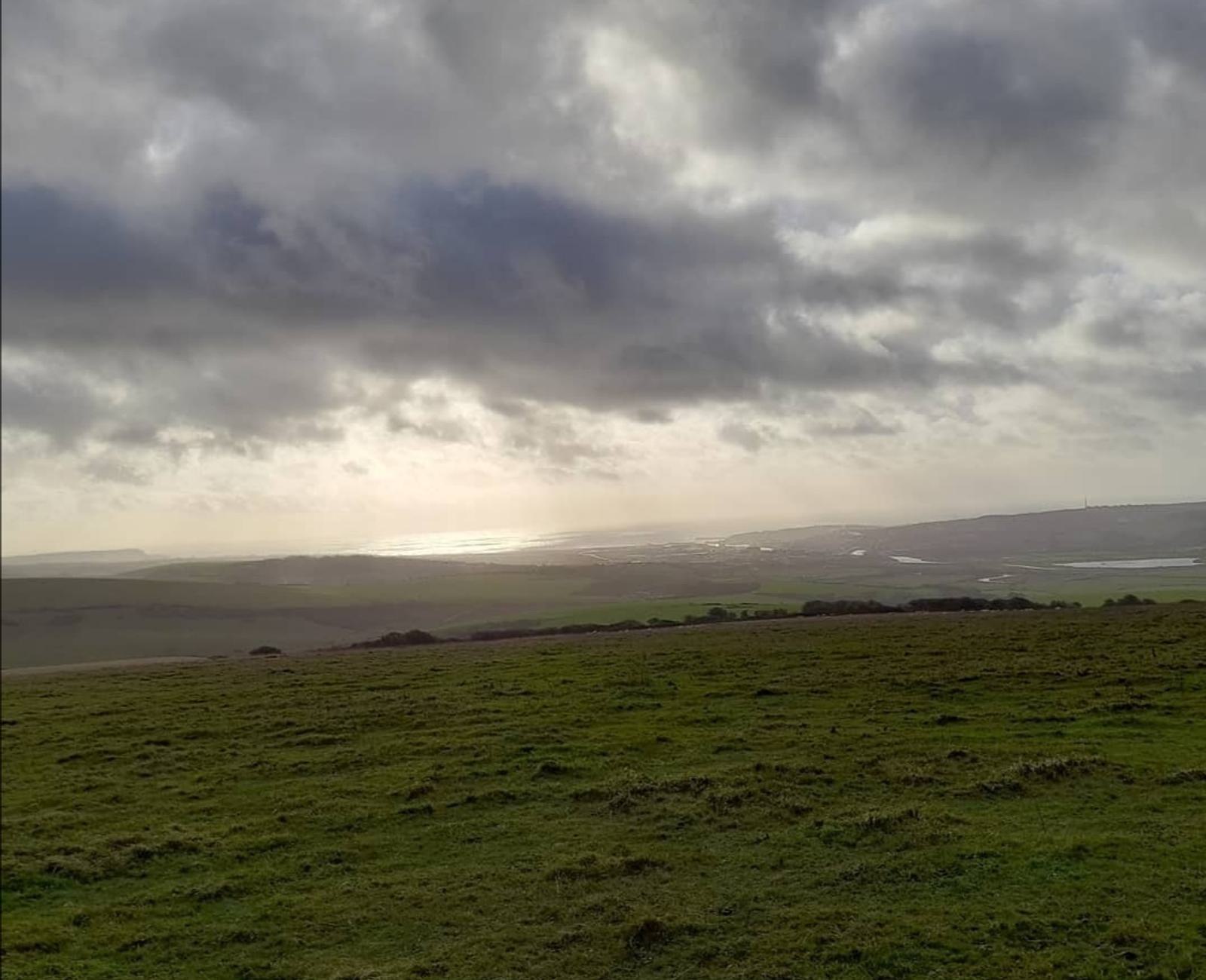 A vast, lush green landscape under a dramatic, cloudy sky with patches of sunlight breaking through. The sun is setting, casting its light on a distant body of water, creating a serene and tranquil atmosphere.