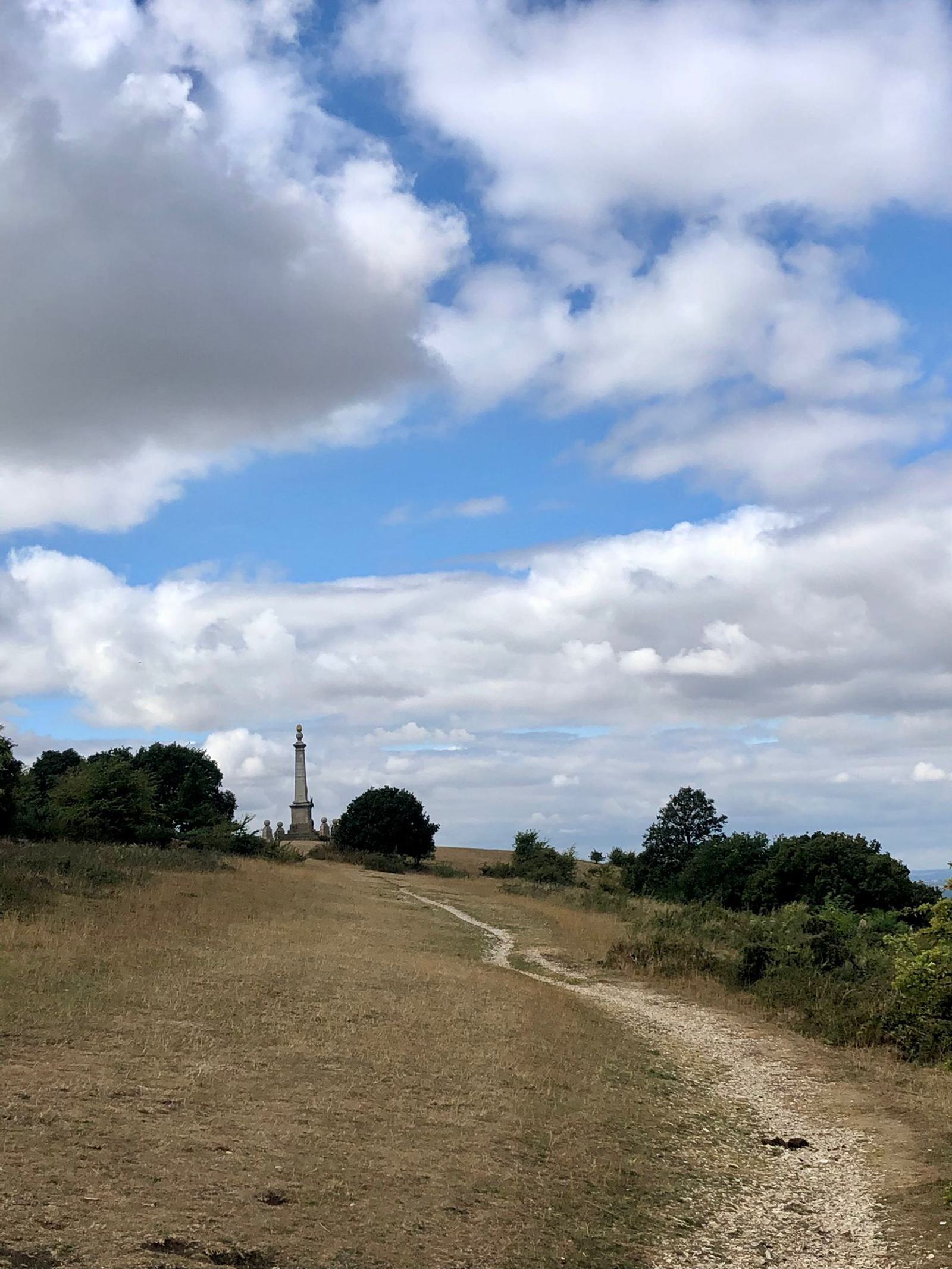 A winding dirt path leads to a lighthouse in the distance, surrounded by a grassy field and scattered trees. The sky above is partly cloudy, with patches of blue visible.
