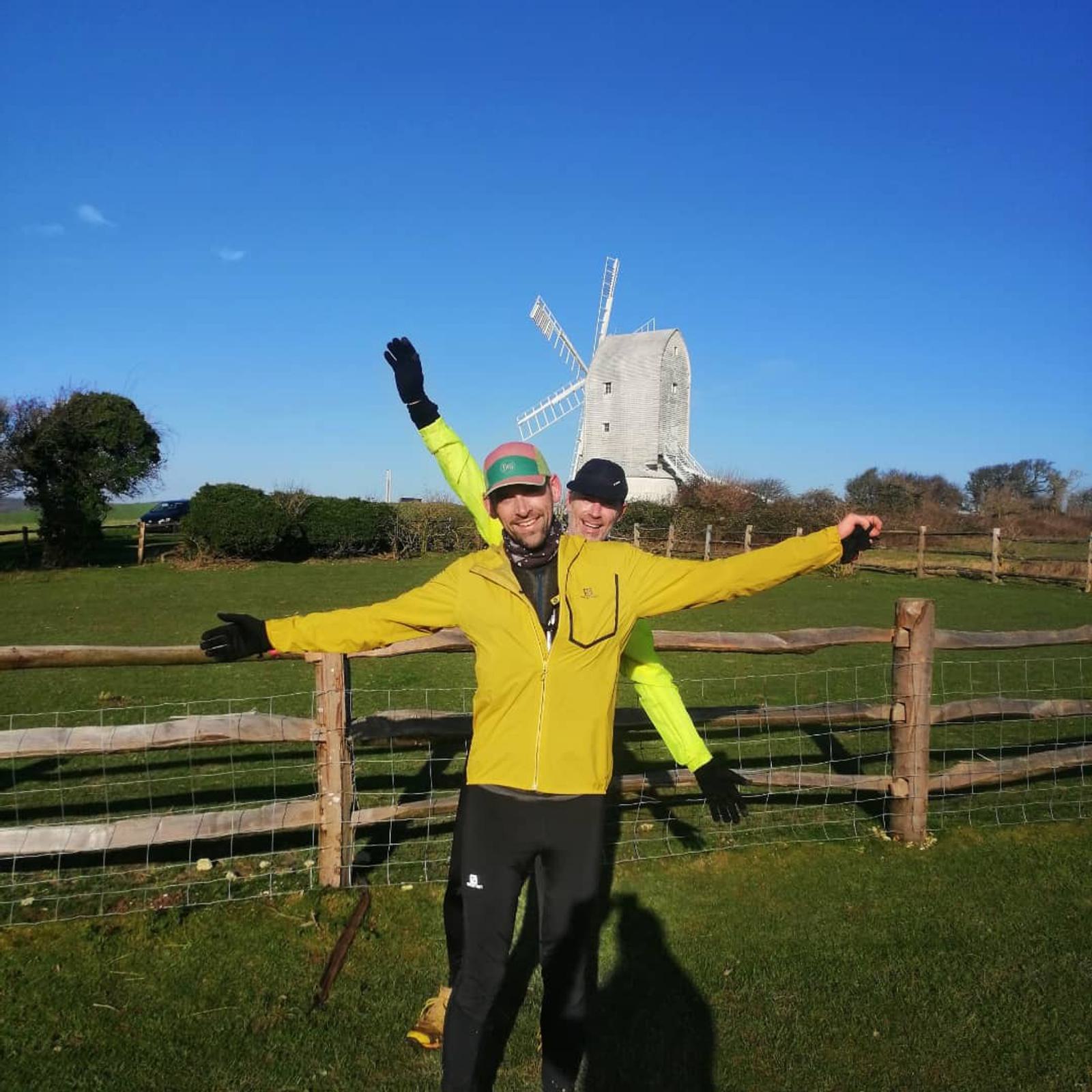Two people wearing brightly colored jackets stand with arms outstretched, one in front of the other, near a wooden fence on a sunny day. Behind them, a large, white windmill and clear blue sky are visible. Both appear happy and are enjoying an outdoor activity.