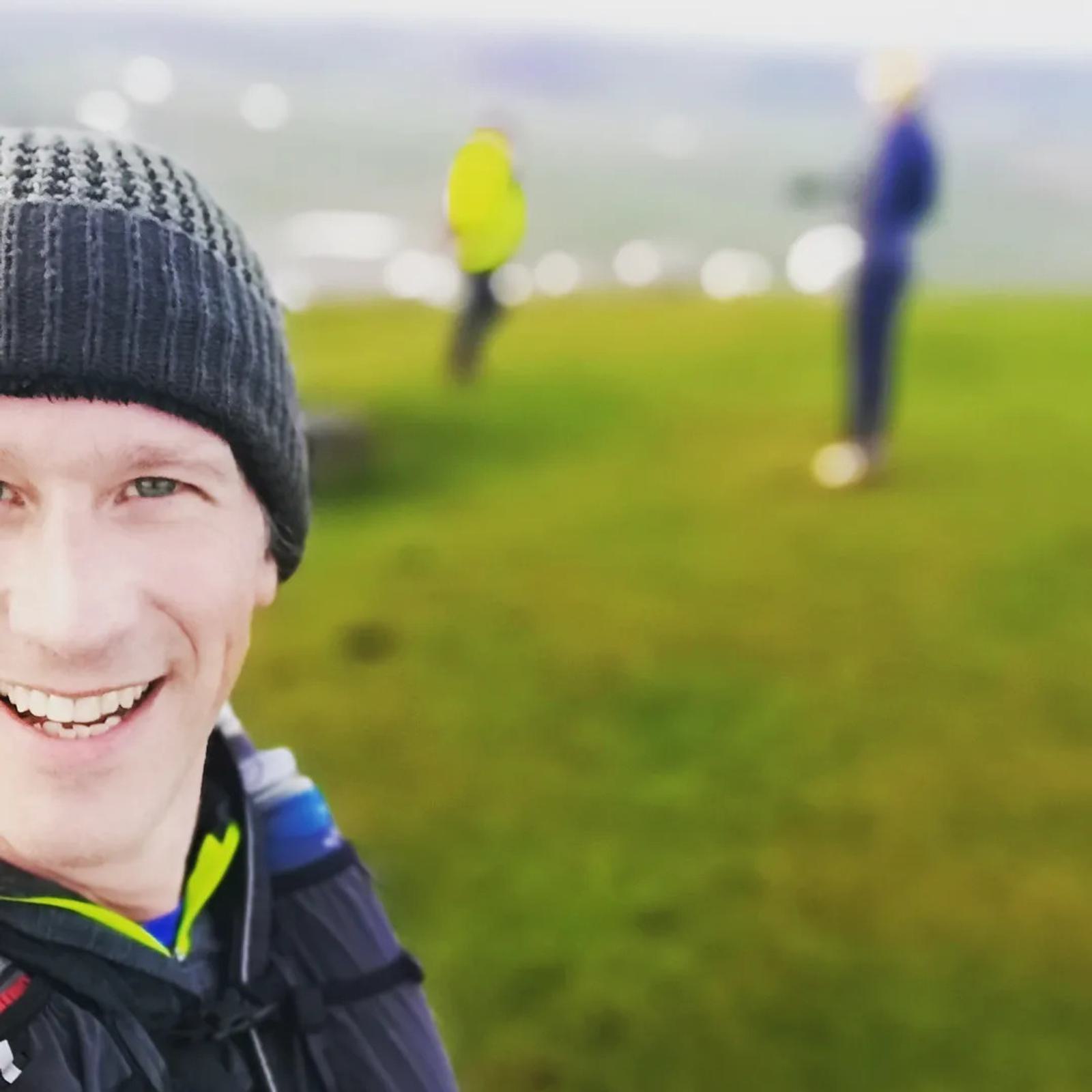 A person wearing a beanie and outdoor clothing smiles at the camera while standing on a grassy hilltop. Two figures in bright jackets stand in the background, slightly out of focus. The scene is set in a scenic, elevated area with blurred, distant landscapes.