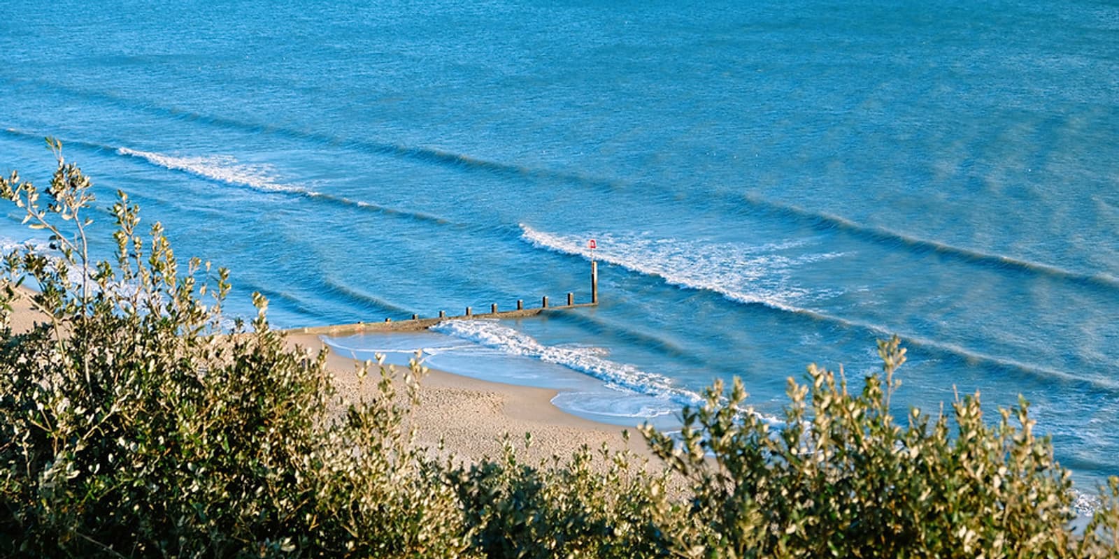 A tranquil beach scene with gentle waves lapping against a sandy shore. A wooden groyne extends into the blue ocean, with green foliage framing the foreground. A small red flag is positioned at the end of the groyne. The sky is clear and blue.